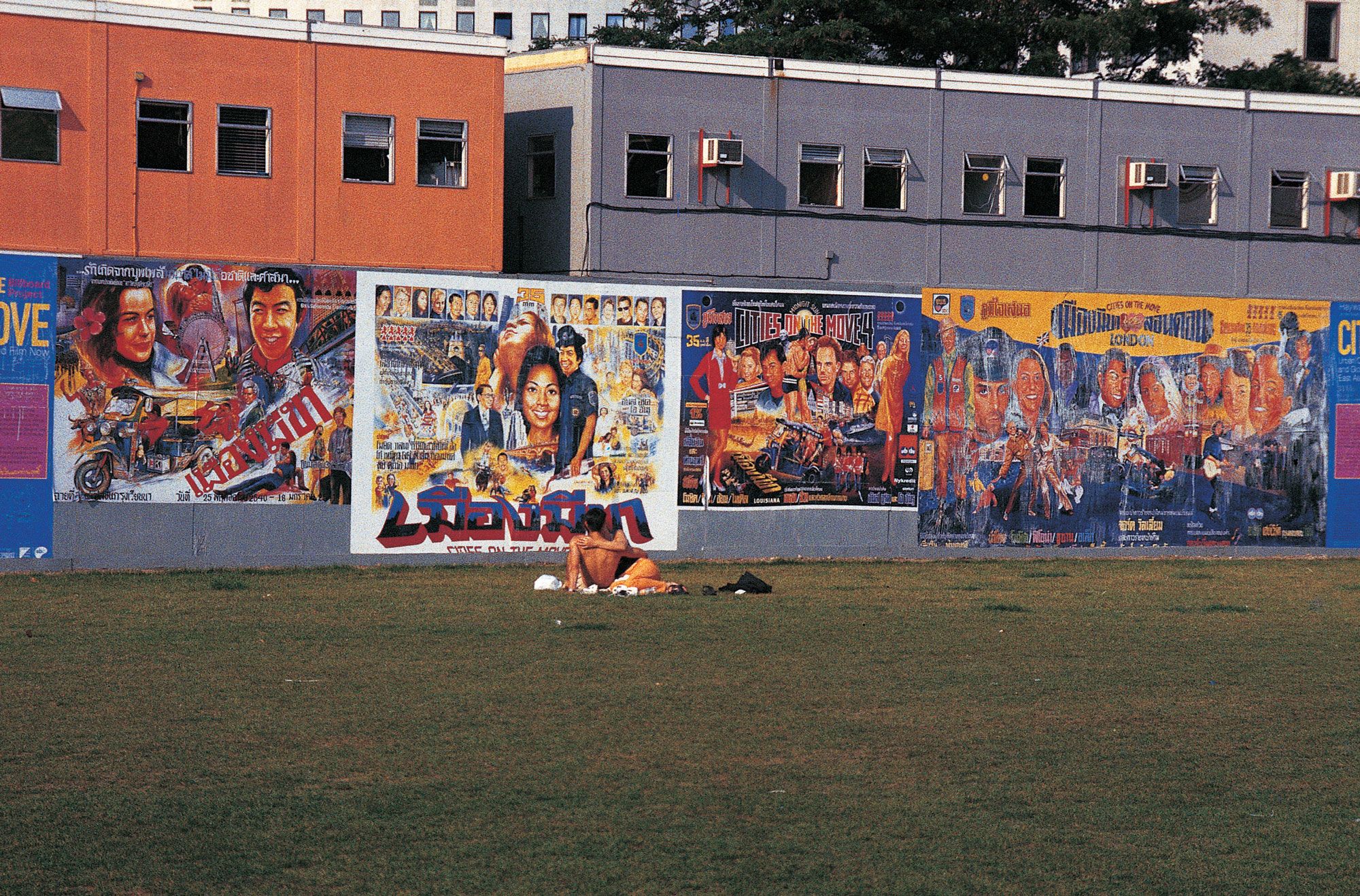 An expanse of lawn stretches before an orange and grey building. Along the edge of the lawn, a hoarding displays multiple colourful posters promoting the ‘Cities on the Move’ exhibition. Two figures lie on the lawn in front of the hoardings, embracing.