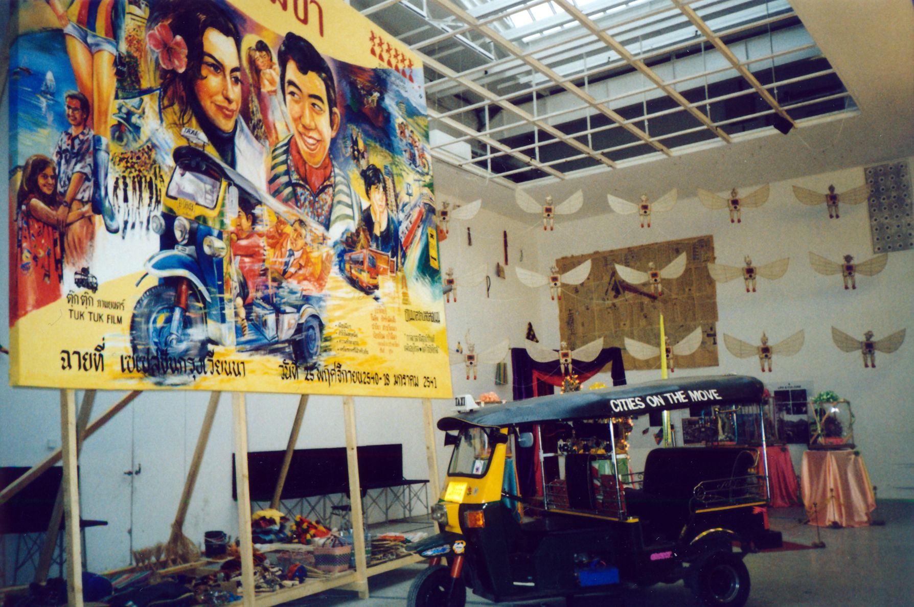 A gallery space showcasing a tuk-tuk and a ‘Cities on the Move’ promotional billboard elevated on wooden planks. The room also features an installation of armless fibreglass figures with insect wings and Javanese-style puppet faces suspended from the ceiling.