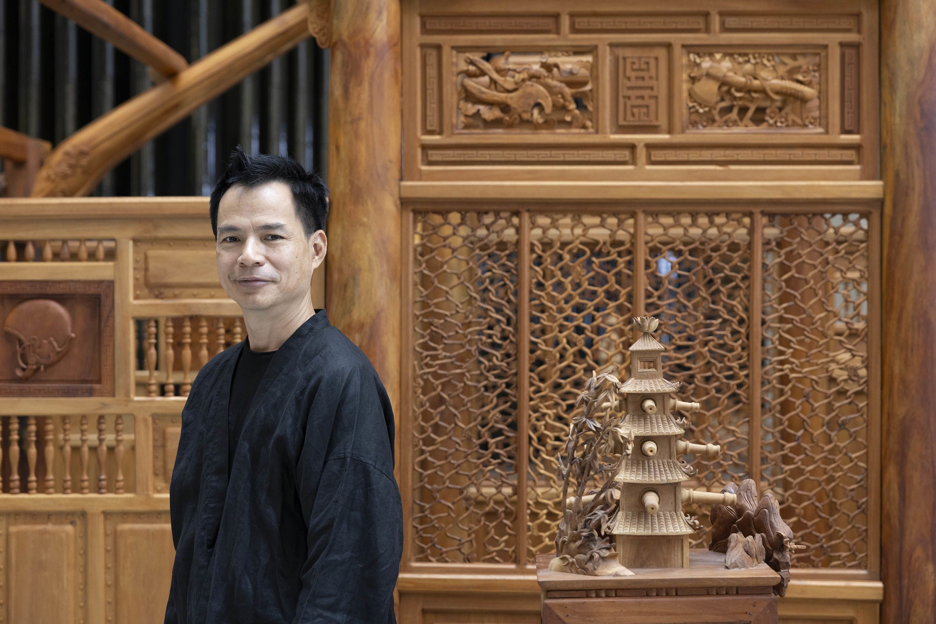 The artist Bùi Công Khánh, dressed in black, stands in front of his wooden sculptural work titled ‘Disolocate’. On our right, a freestanding pagoda sculpture is visible next to the artist. The intricate latticework and reliefs of the main sculpture are visible in the background behind the artist.