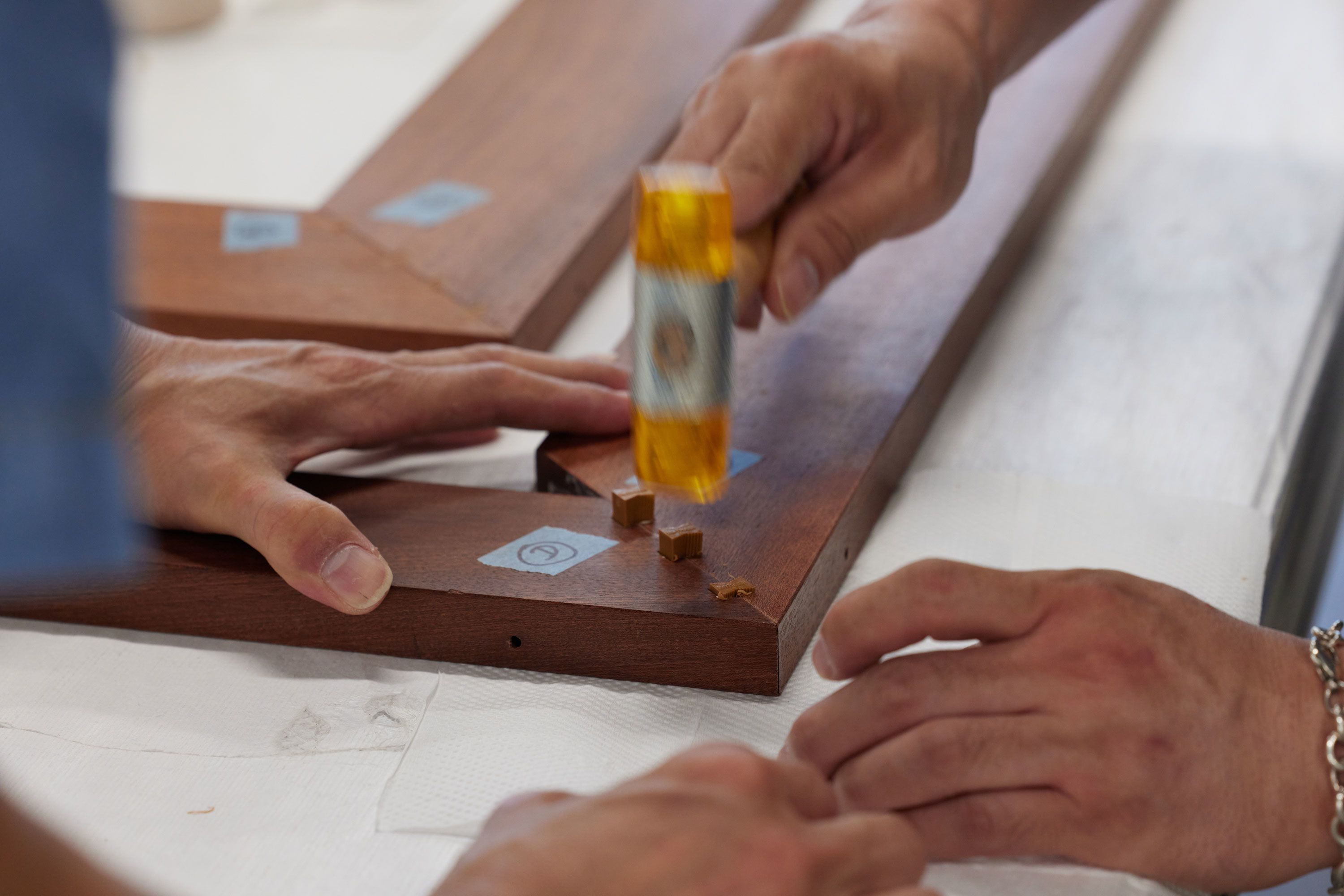 Two pairs of hands are visible, working on the corner of a wooden frame. One pair steadies the pieces of the frame while the other uses a hammer to drive the tenon securely into place.