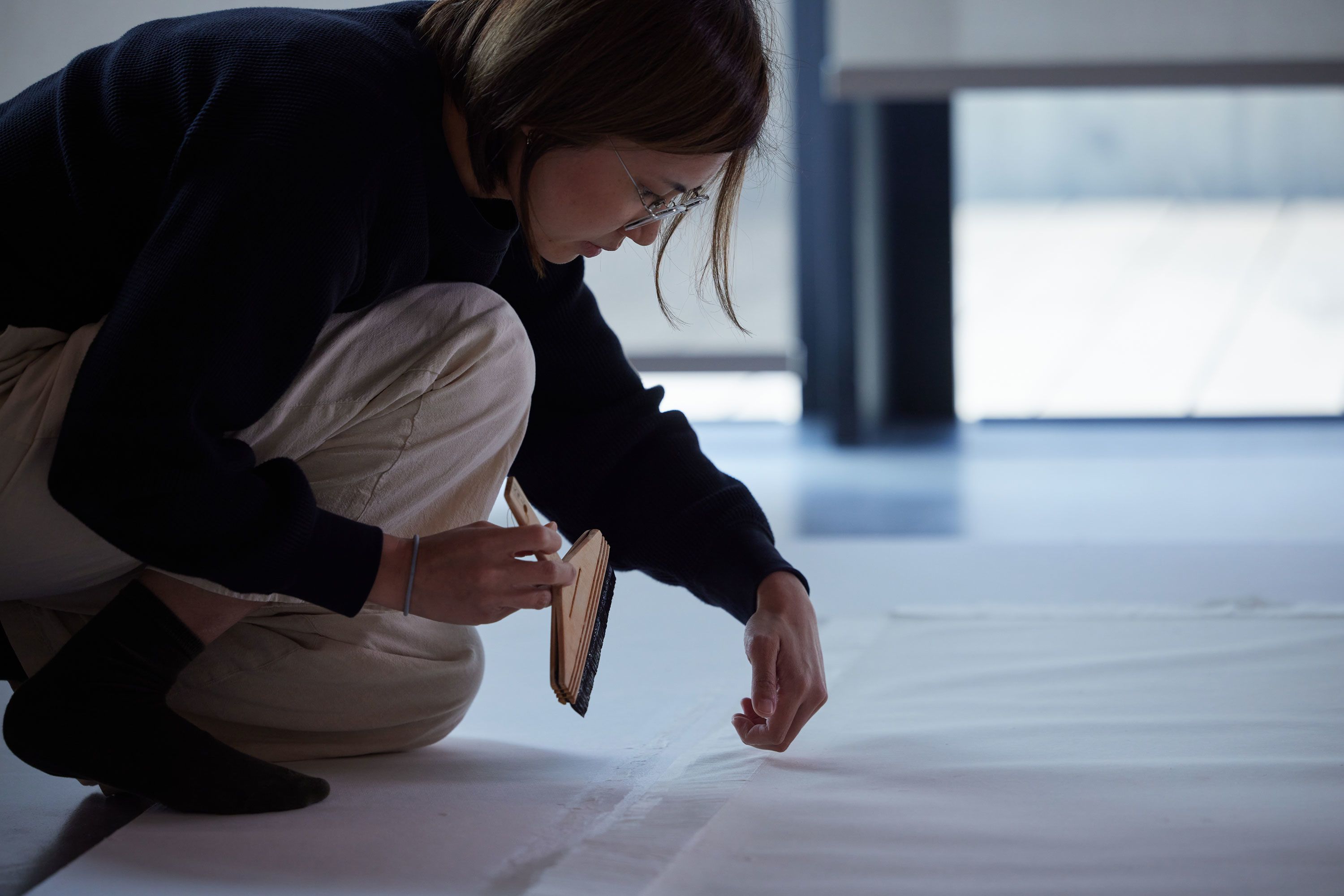 A woman, perched above an artwork lying flat on the ground, holds a broad, soft-bristled flat brush while examining the edge of the work.