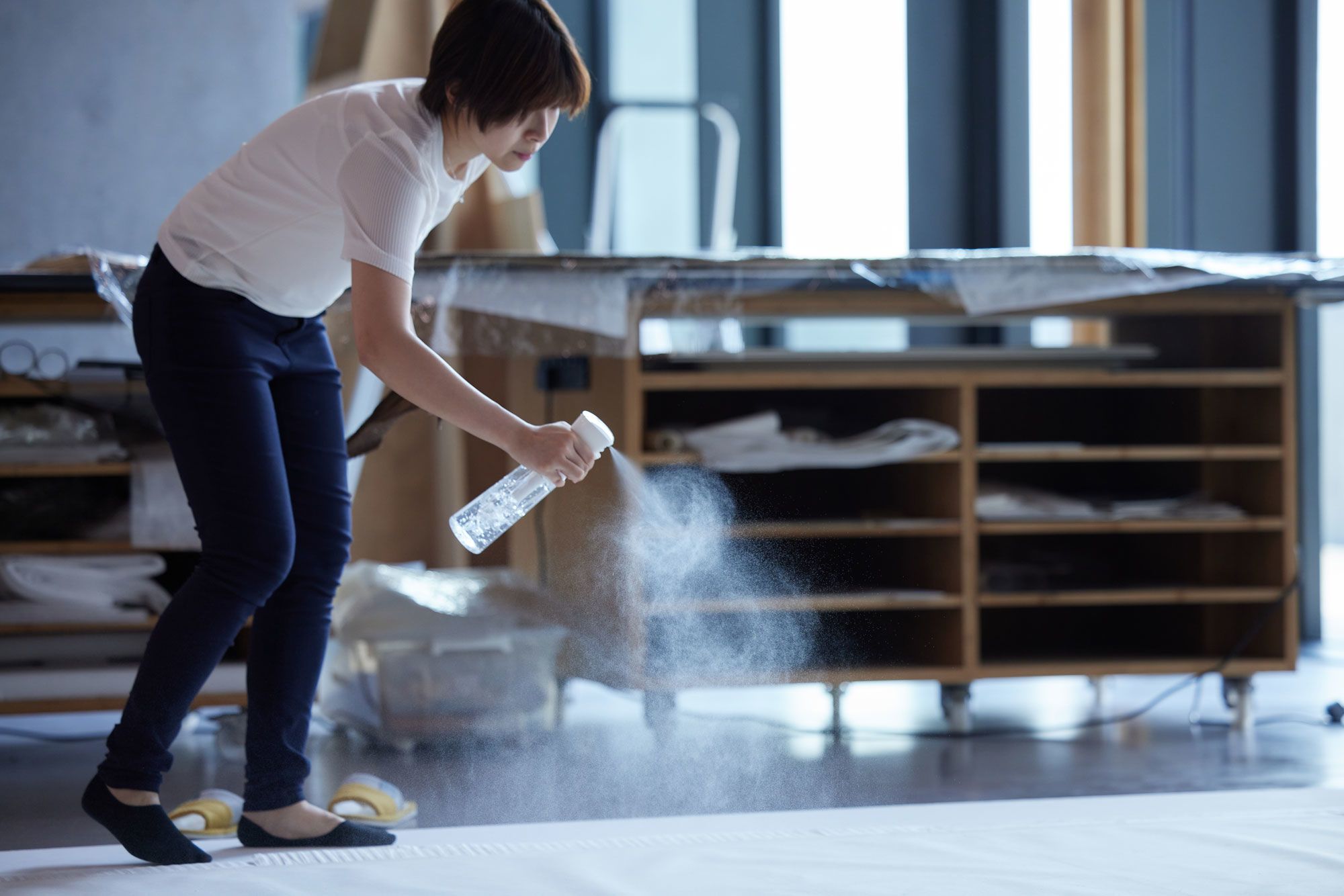Standing in socks, a conservator carefully sprays water as a mist onto an artwork lying flat on the ground. Workbenches are visible in the background.