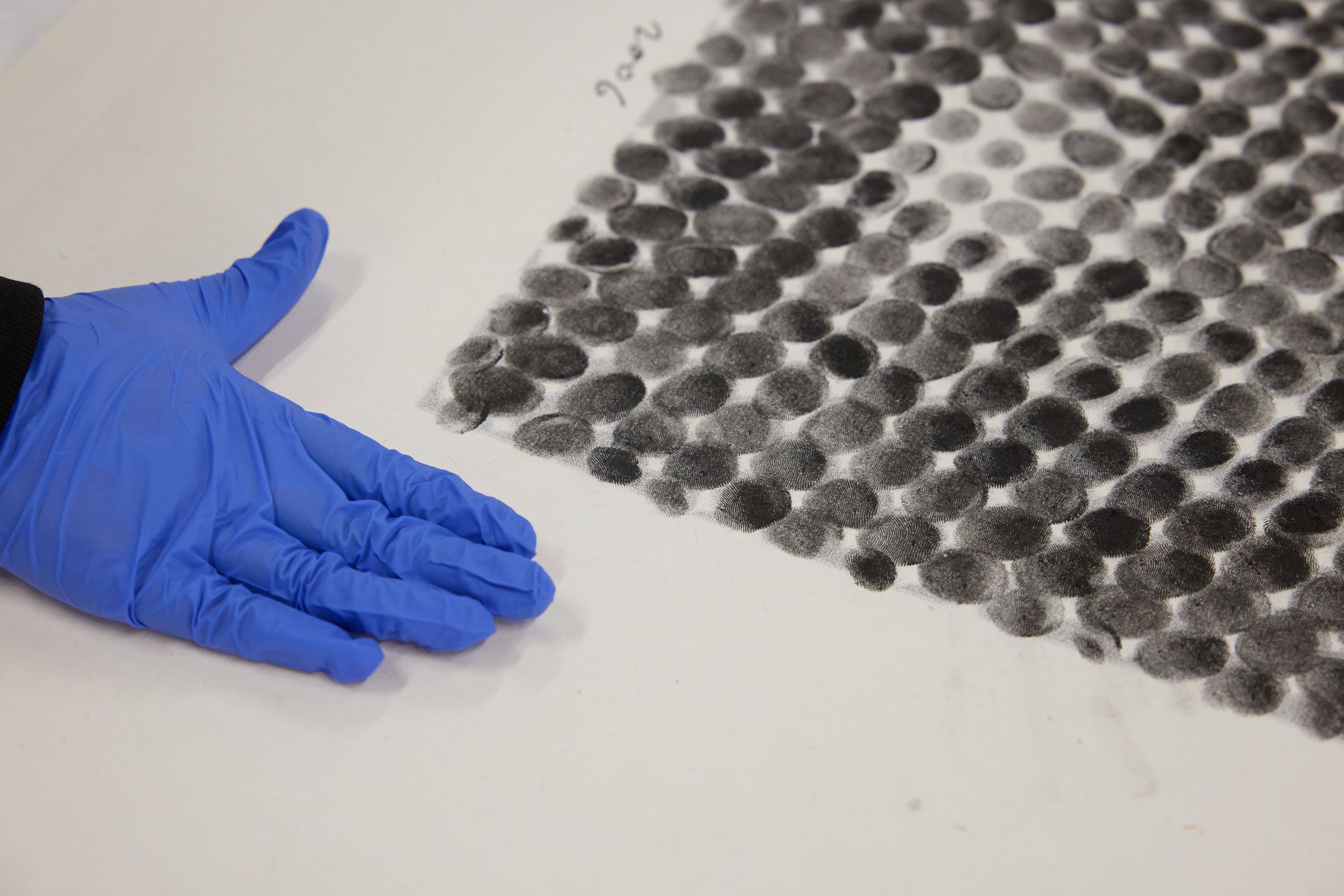 A close-up of an artwork composed of ink fingerprints of varying tonal densities, viewed from the side. A handwritten date, ‘2006’, is visible along the work’s bottom edge. A blue-gloved hand, positioned in a framing gesture, appears in the lower left-hand corner.