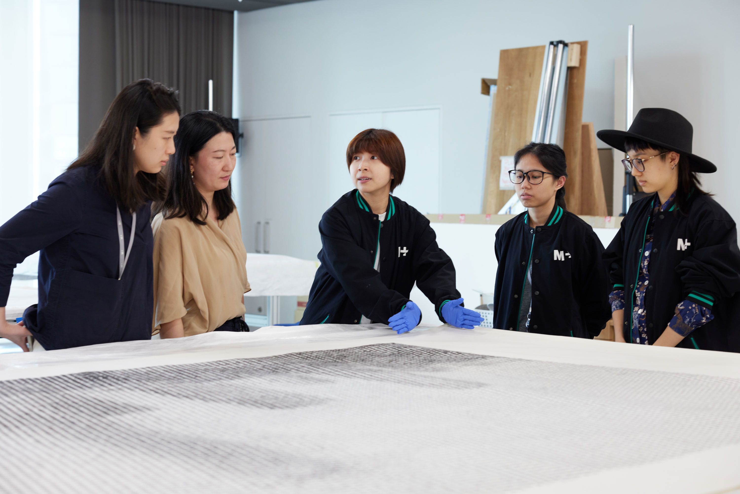 Five women congregate around an artwork laid flat on a table, which takes up the immediate foreground of the image. The woman in the centre of the picture, wearing blue gloves, gestures as she explains something.