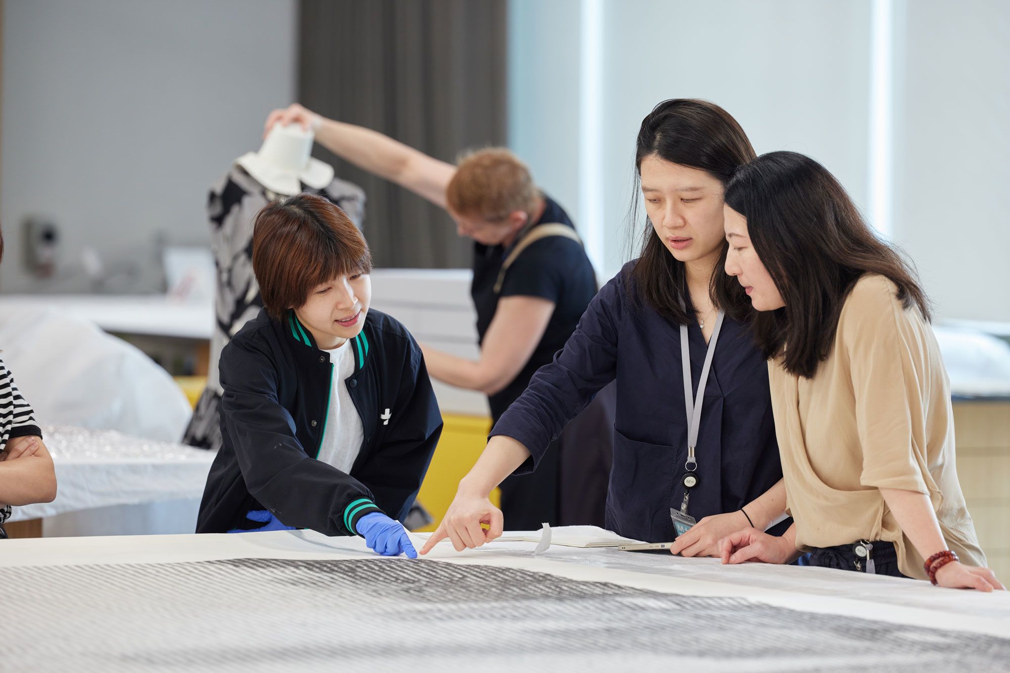 Three women stand around a large-scale artwork laid flat on a table. Two women point towards the bottom right-hand corner of the work, with one wearing blue gloves.