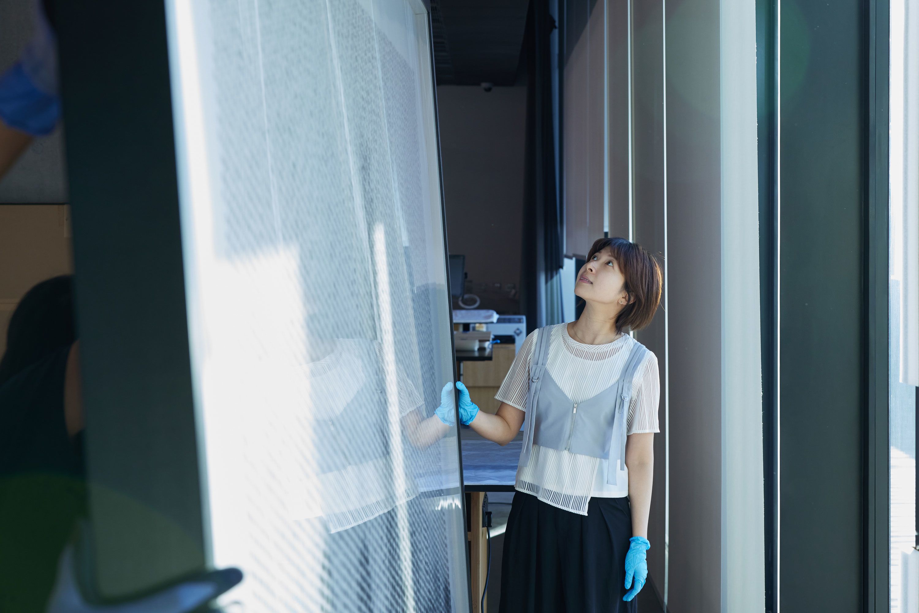 A woman stands at the end of a large framed artwork, looking up at it. She is wearing gloves, and her reflection can be seen in the acrylic surface.