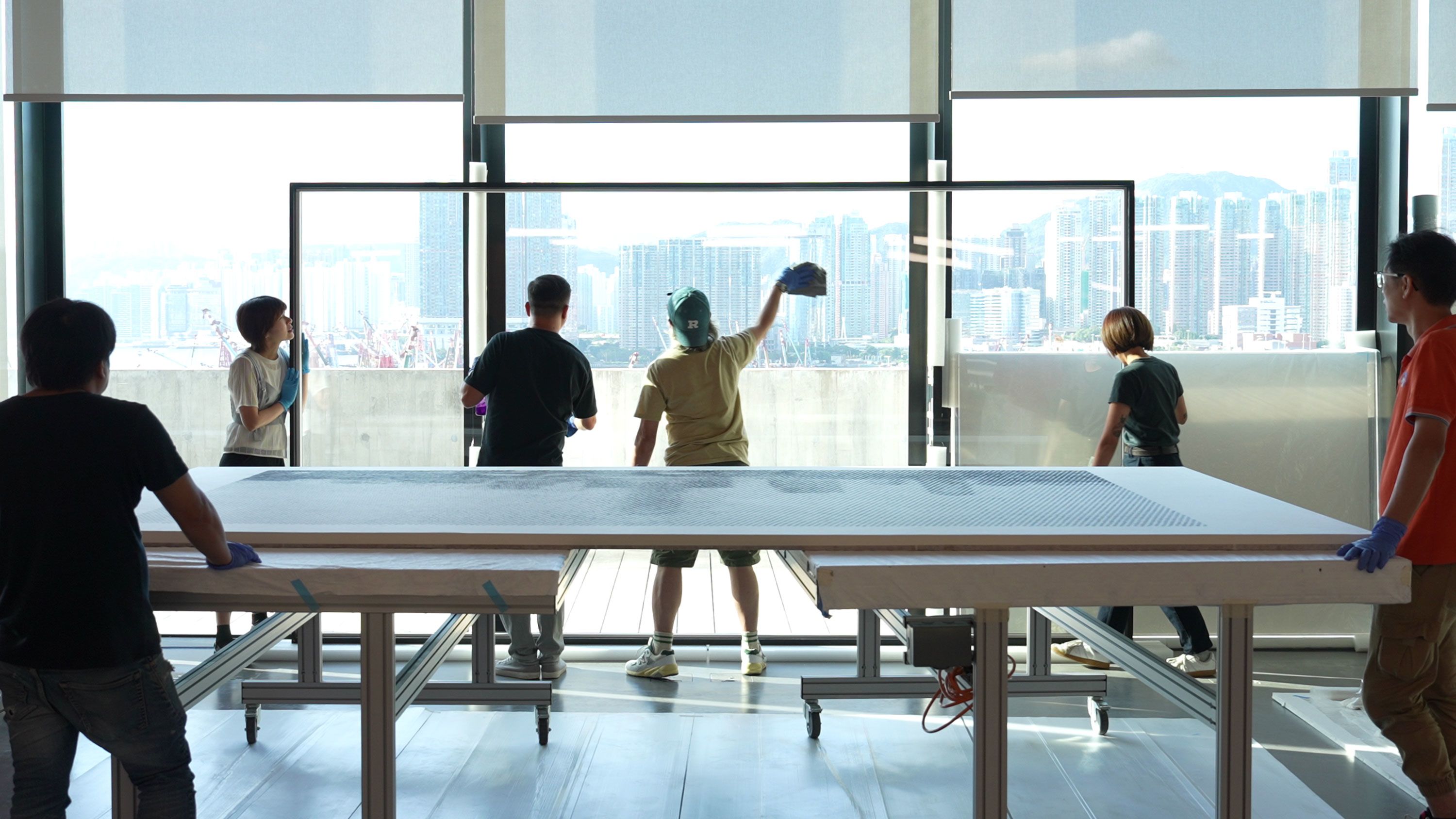 Framers clean a transparent acrylic sheet near floor-to-ceiling windows, which reveals a cityscape. In the foreground, a large work table holds tools and materials for frame assembly.