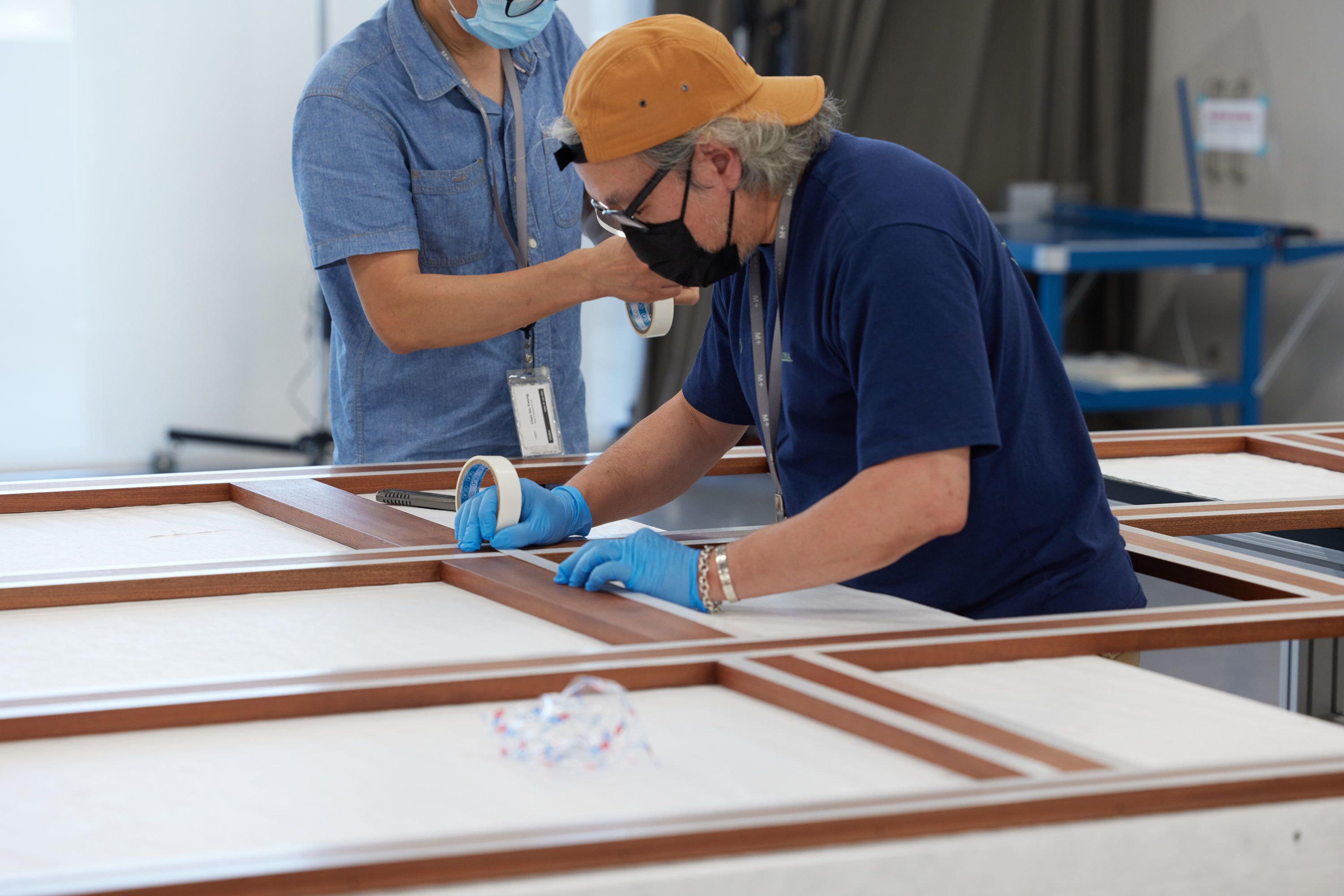 A man wearing a reversed cap stands at the centre of a wooden lattice grid. He carefully applies double-sided adhesive tape along the edges of the lattice structure. 