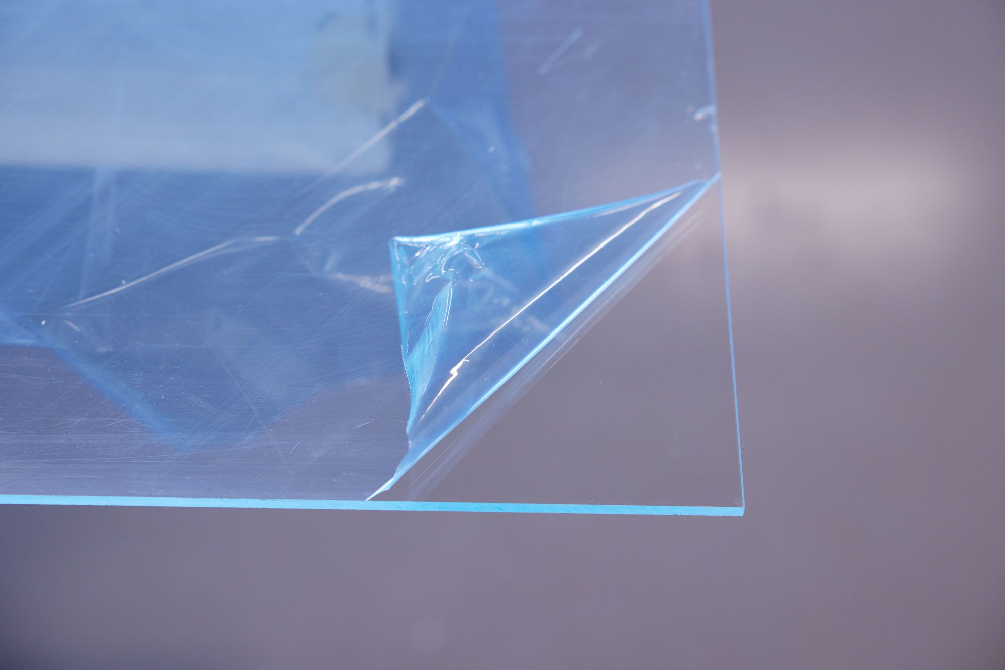 Close-up of a transparent acrylic sheet corner laid flat on a surface. The sheet’s protective plastic wrapping has been partially peeled back in the bottom right corner.