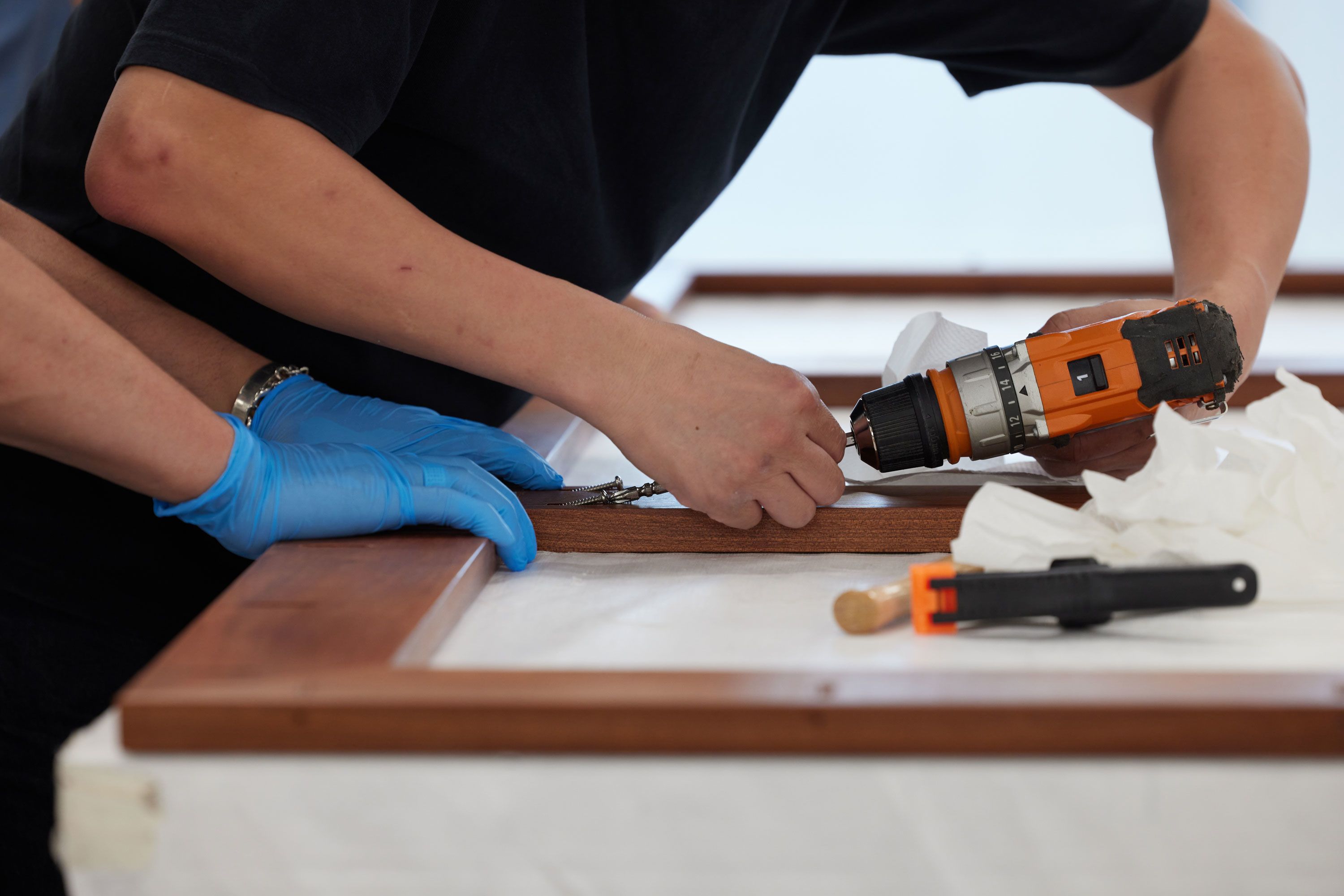 Two pairs of hands work on assembling an artwork’s frame. Two gloved hands firmly hold the wooden pieces in place while the other pair uses a power tool to drill into the frame.