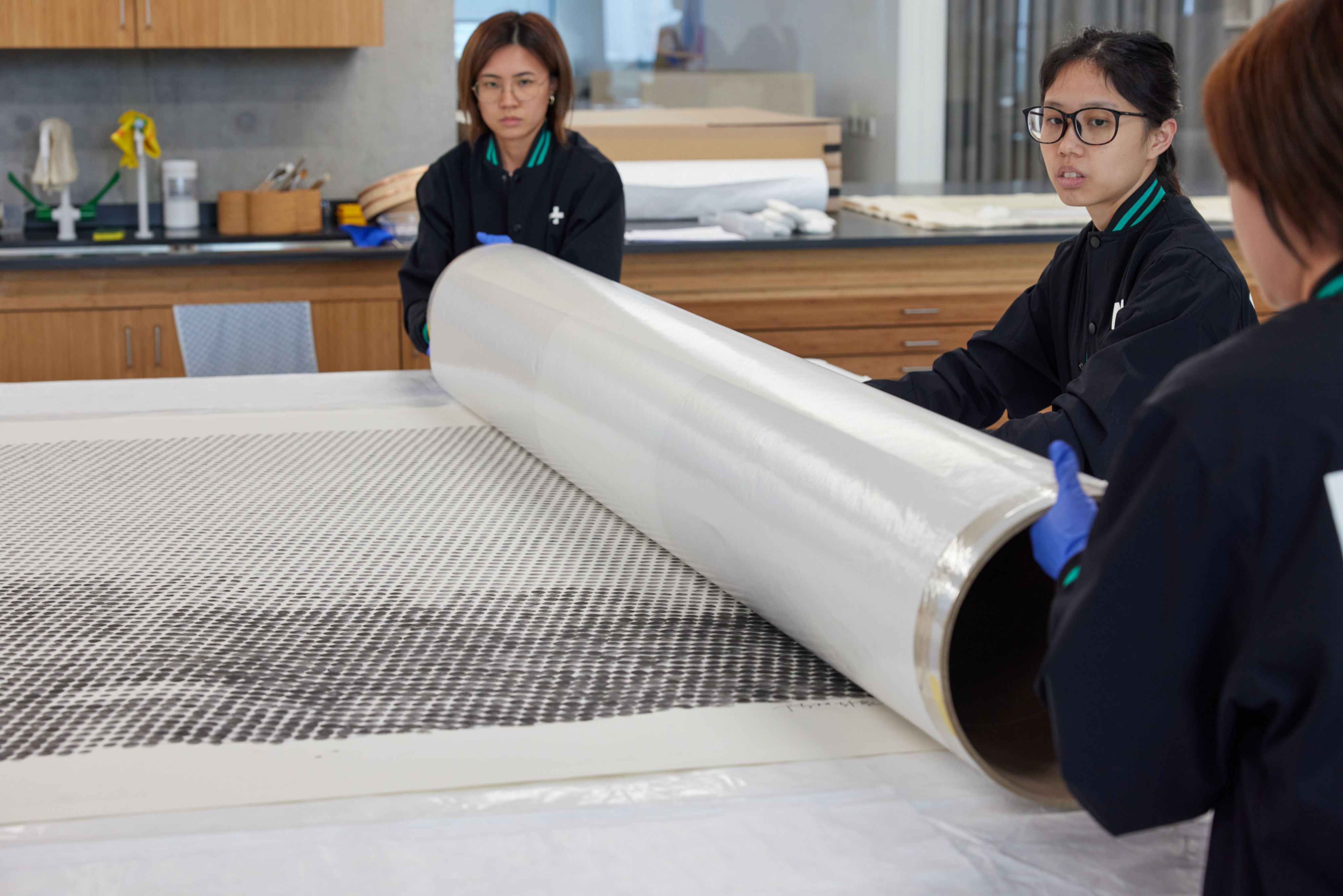 Three women carefully unroll a paper artwork, composed of ink fingerprints of varying tonal densities, onto a large table.