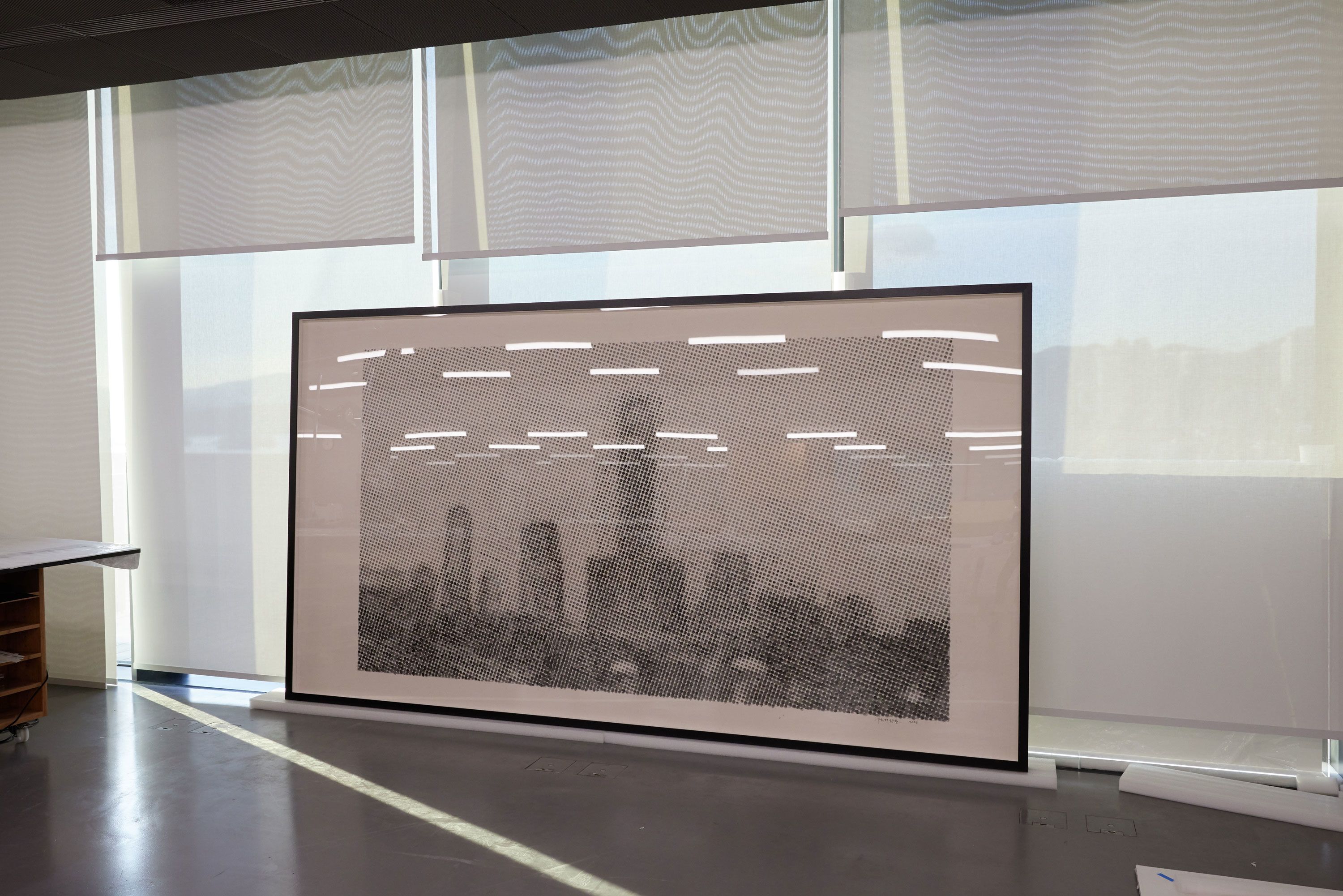 A framed artwork rests on padded plastic sheeting in a conservation studio, leaning against floor-to-ceiling windows partially covered by semi-opaque blinds. Fluorescent lights reflect on the acrylic pane.