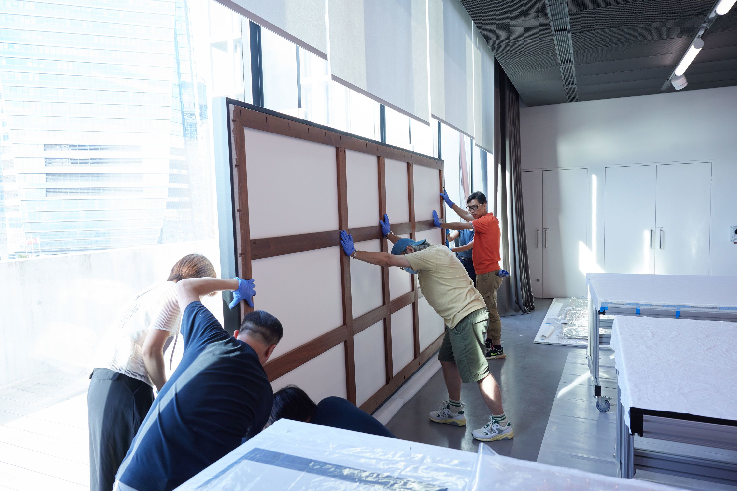 Five people are evenly spaced along the backside of an artwork’s frame, collaboratively affixing it to a backing board holding the mounted artwork.
