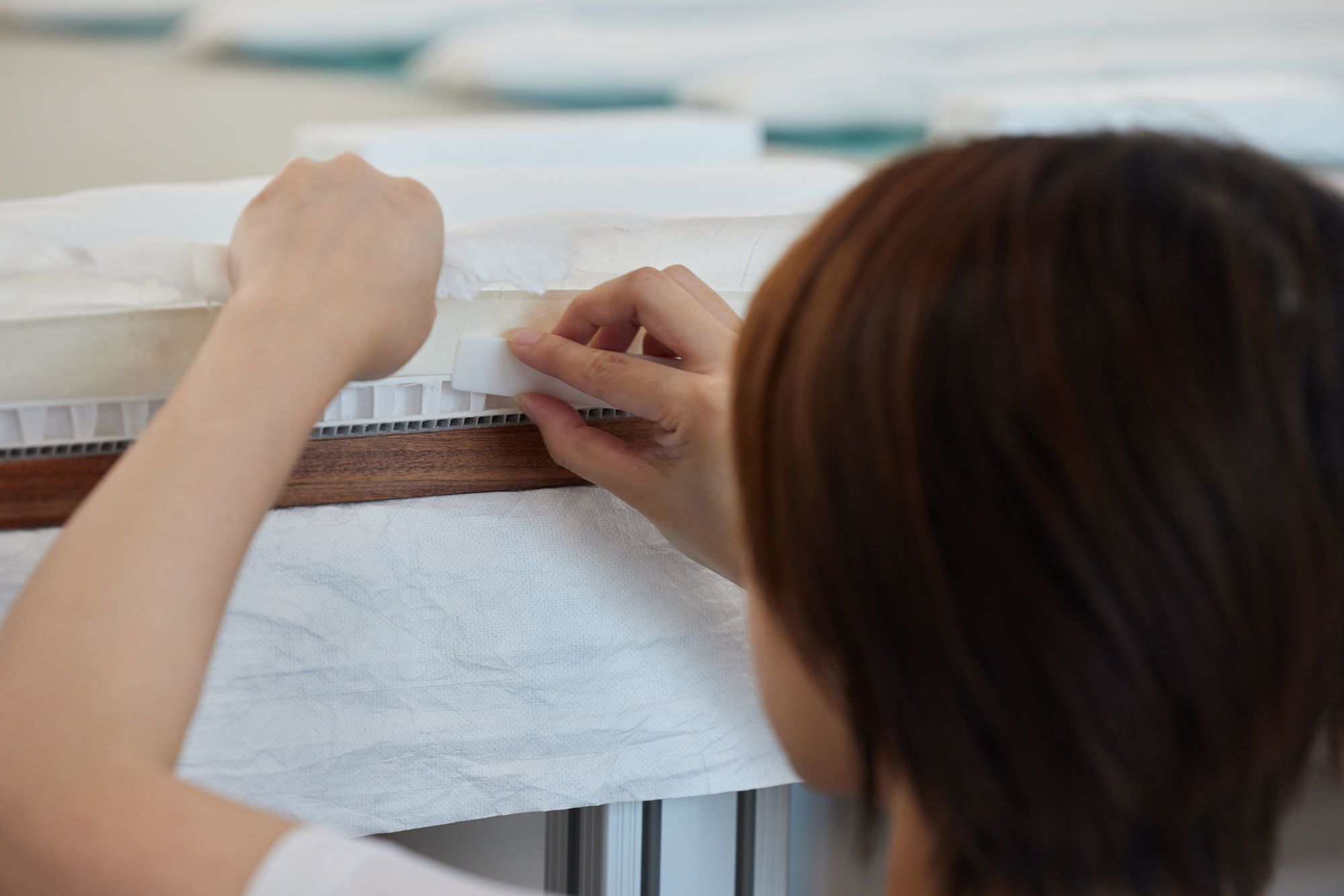 A person, seen from behind, uses their hands to carefully press the edges of thin paper strips onto the surface of a cotton rag mat board.