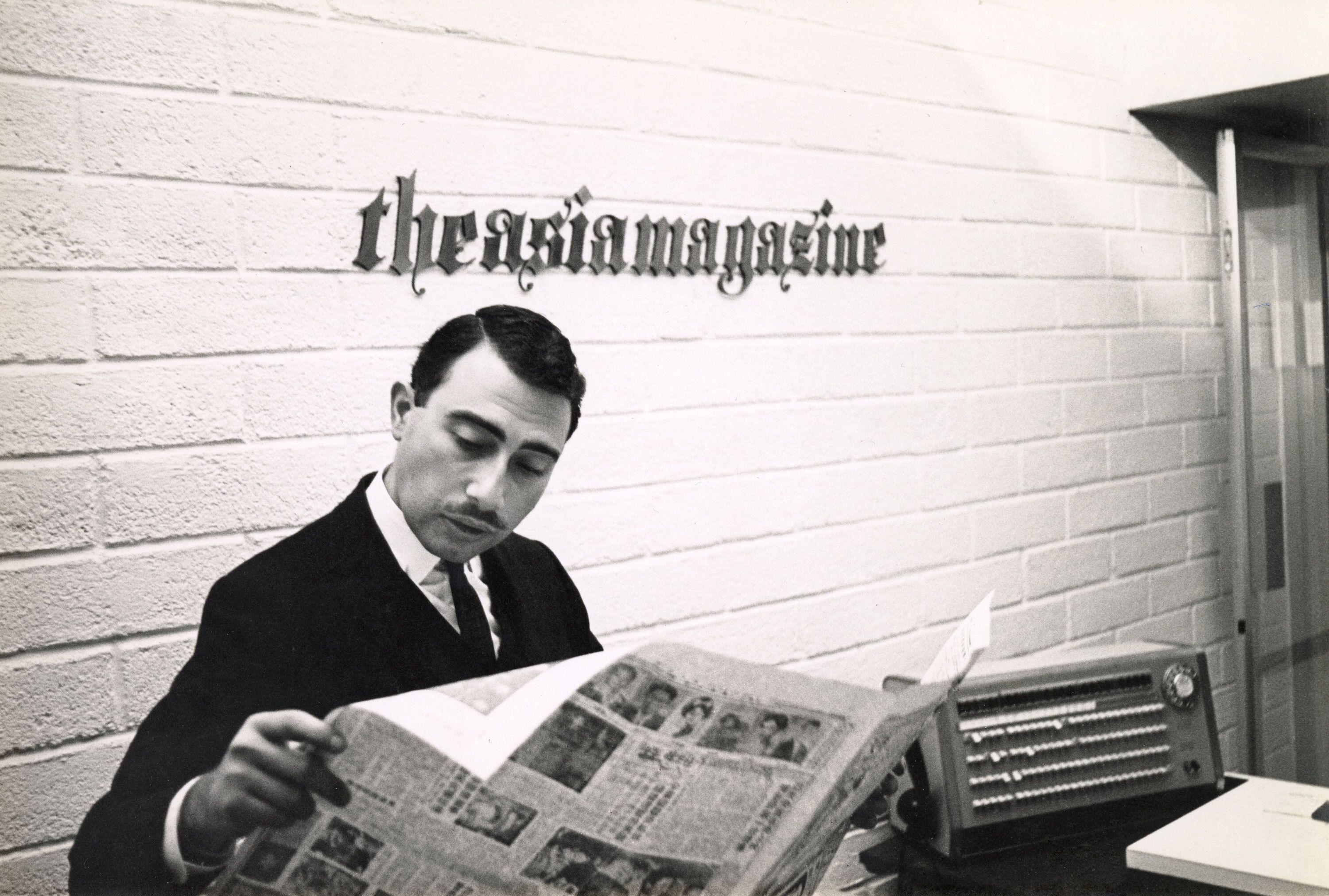 Monochrome photograph of a man in a suit reading a newspaper at a desk, with the words ‘the asia magazine" displayed on the wall behind him.