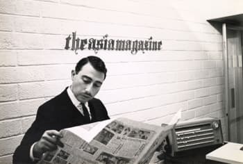 Monochrome photograph of a man in a suit reading a newspaper at a desk, with the words ‘the asia magazine" displayed on the wall behind him.