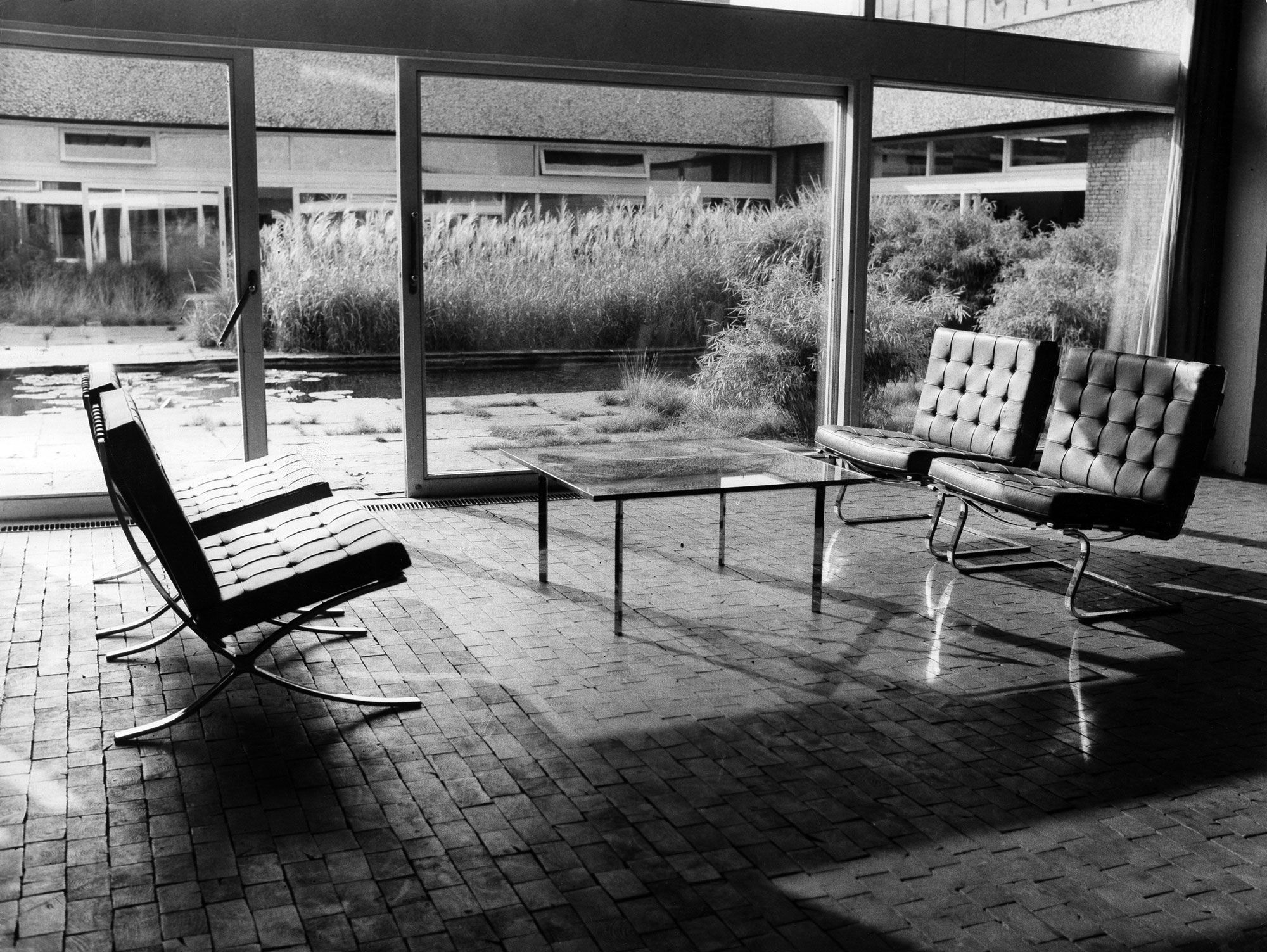 Monochrome photograph of a living room with large glass windows, and four leather and steel modernist chairs around a glass table. Outside lies a garden and a pond.