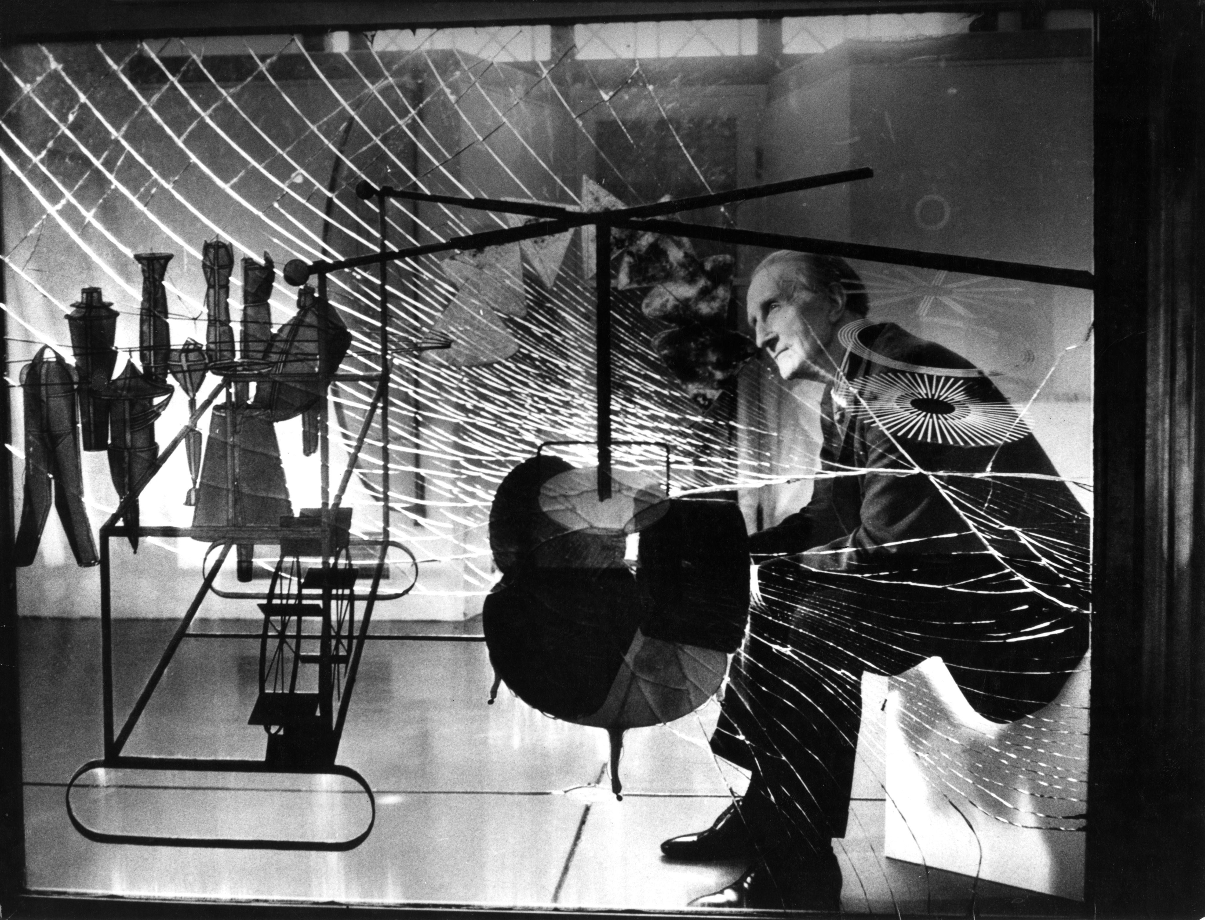 A monochrome image shows Marcel Duchamp, dressed in a suit, seated behind an artwork of fractured glass. Geometric shapes within the glass form a mechanical assemblage, strikingly framing the entire scene.