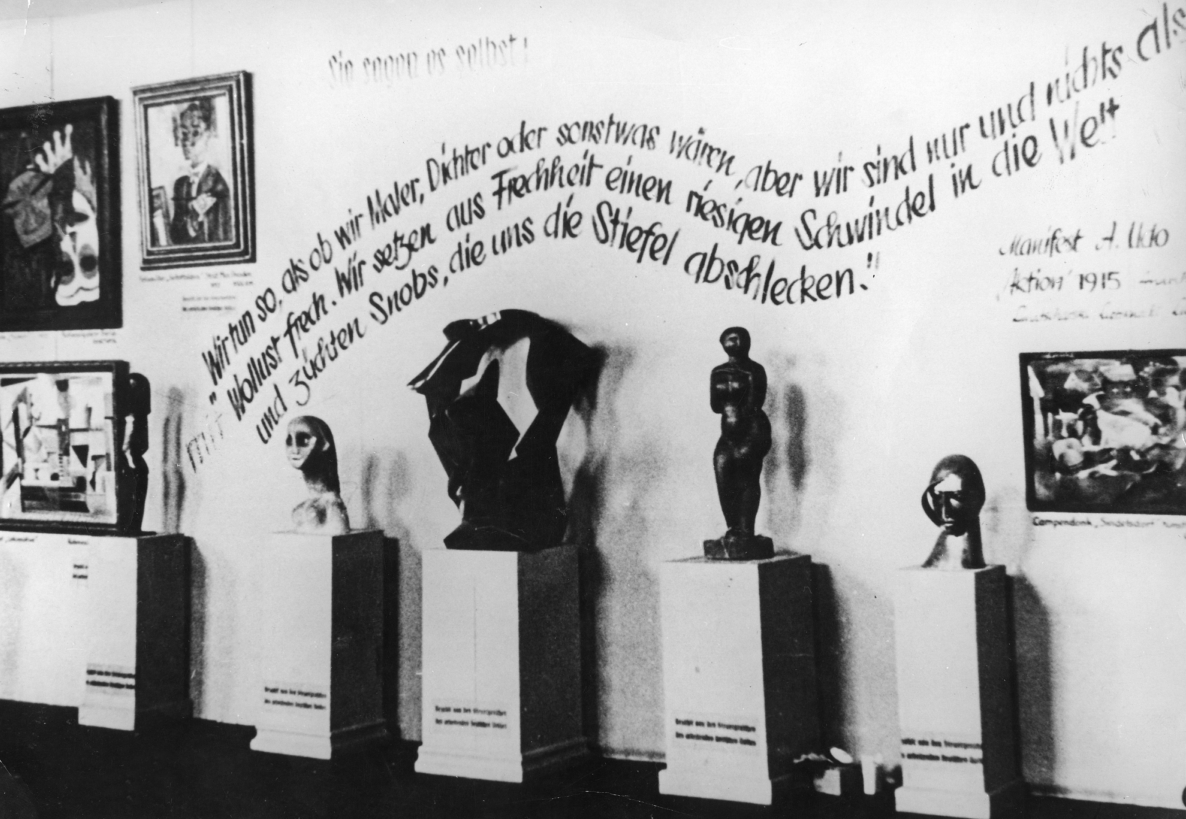 Monochrome photograph shows five abstract human-like sculptures on pillars in a gallery. German text on the wall behind them reads: ‘We pretend to be painters, poets, or to have meaning, but we are nothing but impertinent deviants. We deliberately perpetuate a great fraud into the world and breed snobs who lick our boots.’, alongside the paintings.