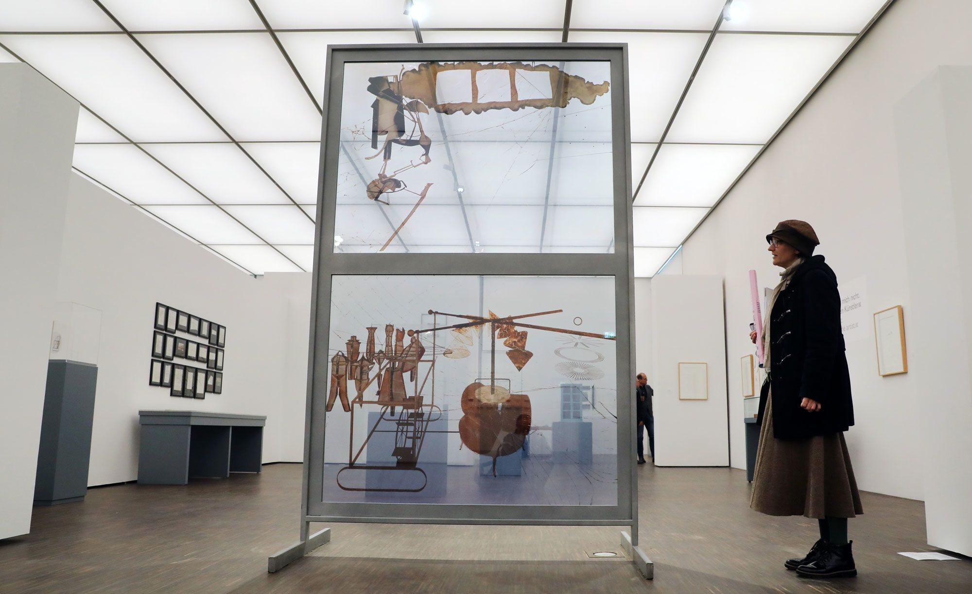 A sculptural work of two vertical glass panes with abstract designs stands in a gallery, their transparent surfaces catching the light from the illuminated roofing above. A visitor observes the work.