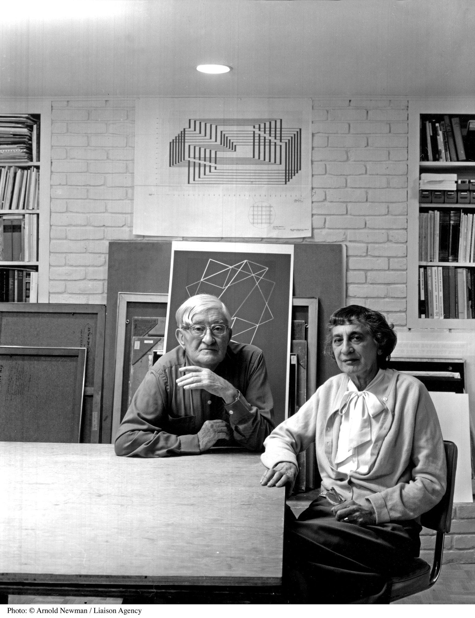 Monochrome photograph of the middle-aged Josef and Anni Albers sitting at a wooden table. Behind them are geometrical drawings propped up against the brick wall, a geometrical drawing on the wall, and two bookshelves on the sides. 