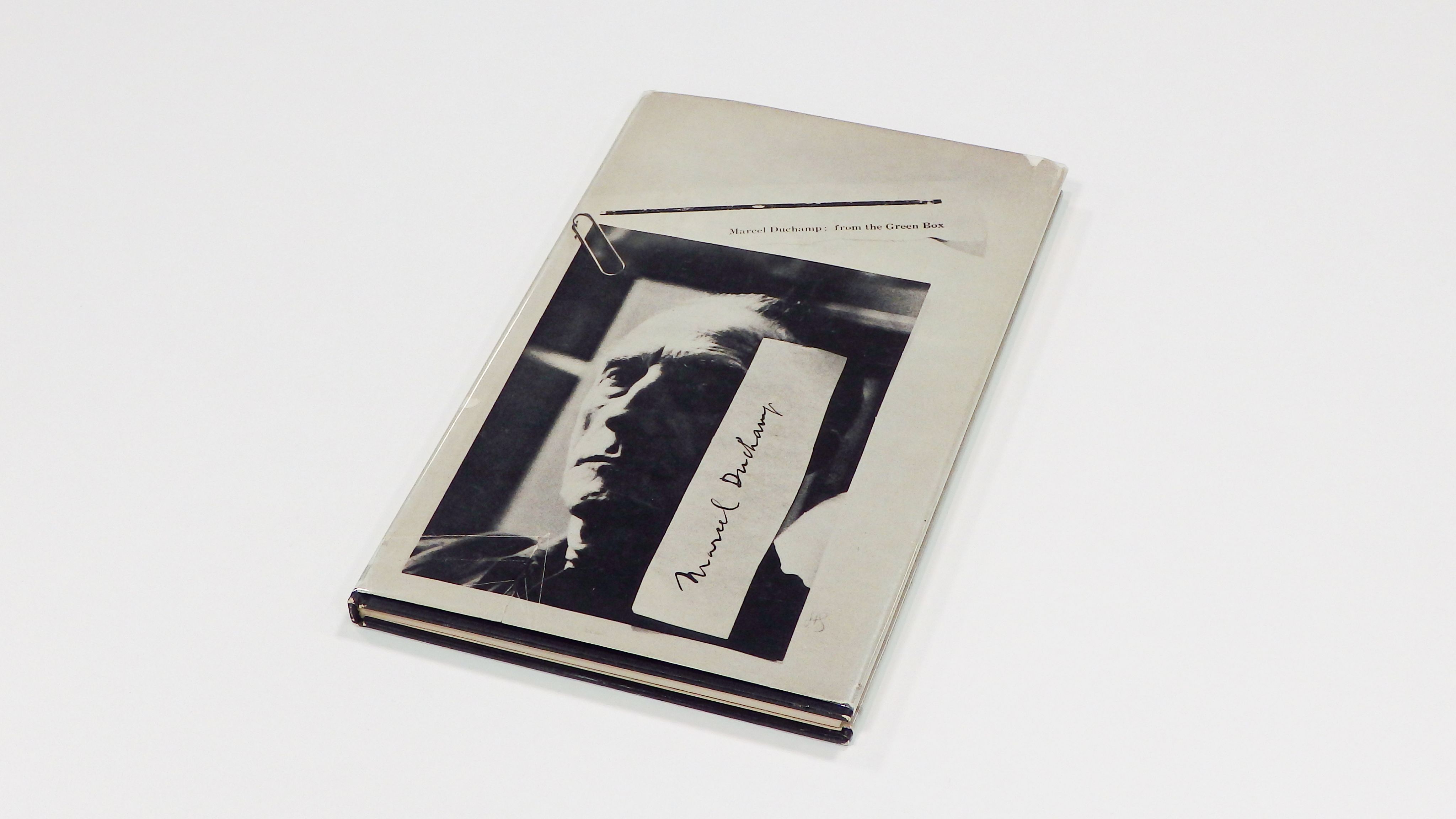A book titled ‘Marcel Duchamp: From the Green Box’ rests on a white surface. The book cover features a black-and-white portrait of Marcel Duchamp, adorned with torn pieces of paper. The initials ‘HS’ are faintly visible beside Duchamp’s image.