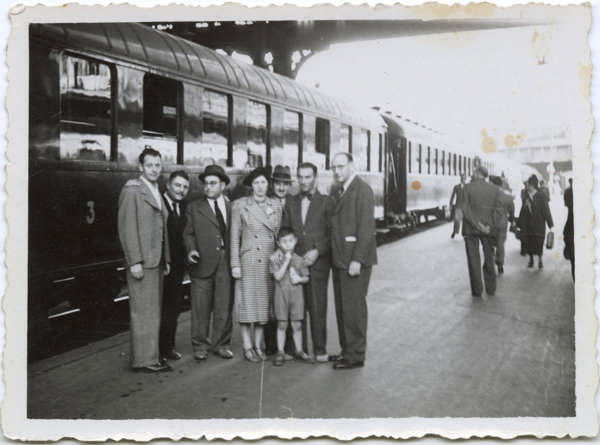 Monochrome photograph depicts eight figures, seven adults and one child, formally dressed on a train station platform. A train looms behind, while commuters pass. Light stains mark the photograph’s surface.