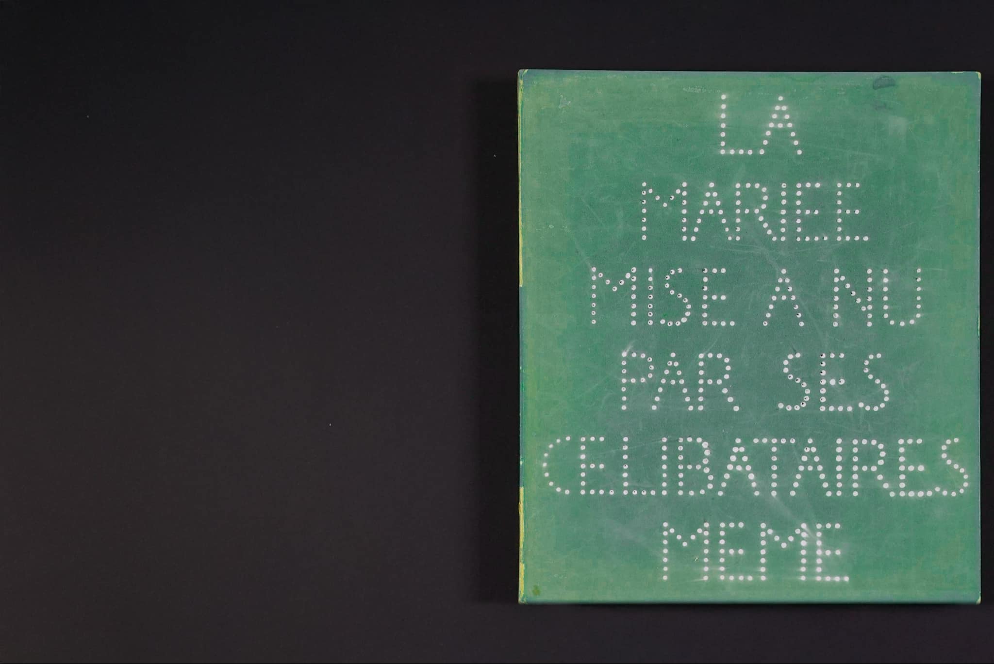 The cover of a green box displays the French title ‘LA MARIEE MISE A NU PAR SES CELIBATAIRES MEME’, written entirely in dots. The text appears bold and textured.