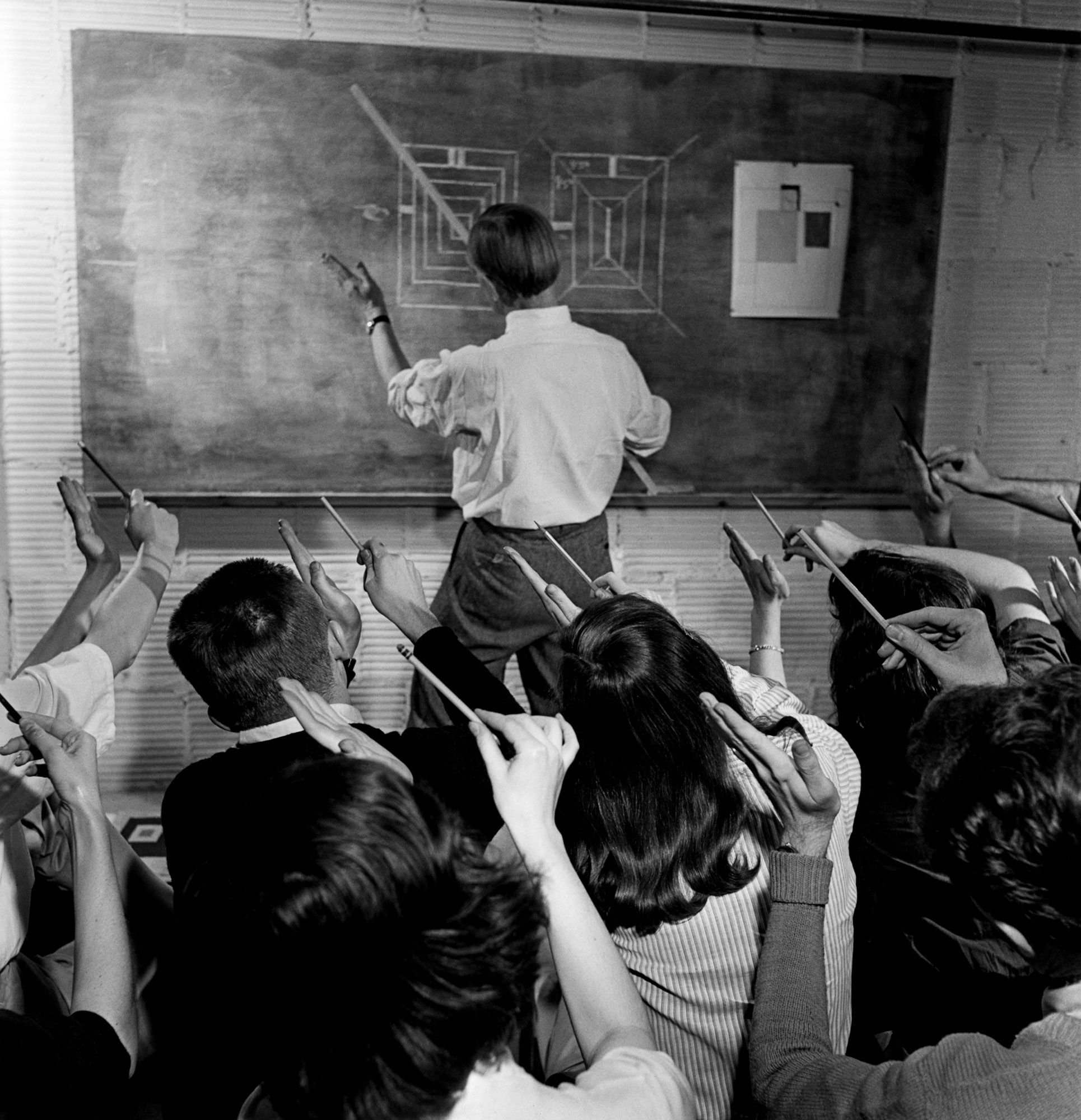 Monochrome photograph of a classroom where a design lecturer stands at the front, holding a ruler at a 45-degree angle to match geometric shapes on the blackboard. His left hand mimics the angle, while students in the foreground, holding pencils, mirror his gesture.