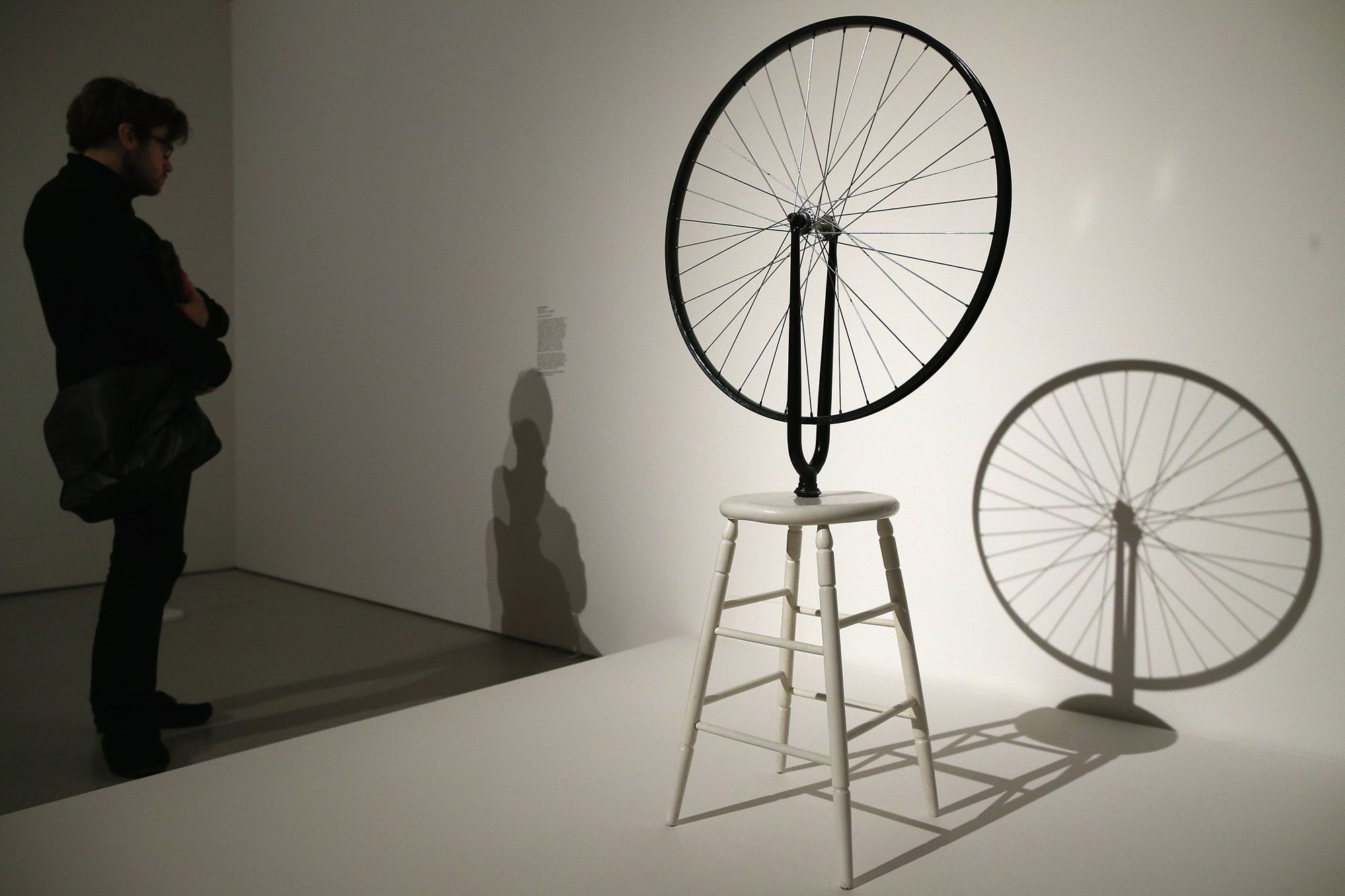 Sculptural artwork of a bicycle wheel on a white stool casts a dramatic shadow on the gallery wall while a visitor reads a wall label beside it.
