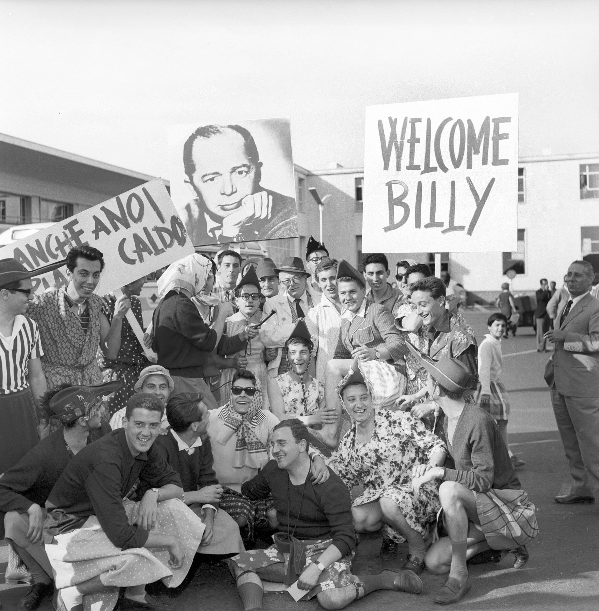 Monochrome photograph of a joyous group outdoors surrounding Billy Wilder, all dressed in robe-like clothing, smiling and posing. One person holds a ‘WELCOME BILLY’ sign, another holds a portrait, and a third displays partially obscured sign in Italian.