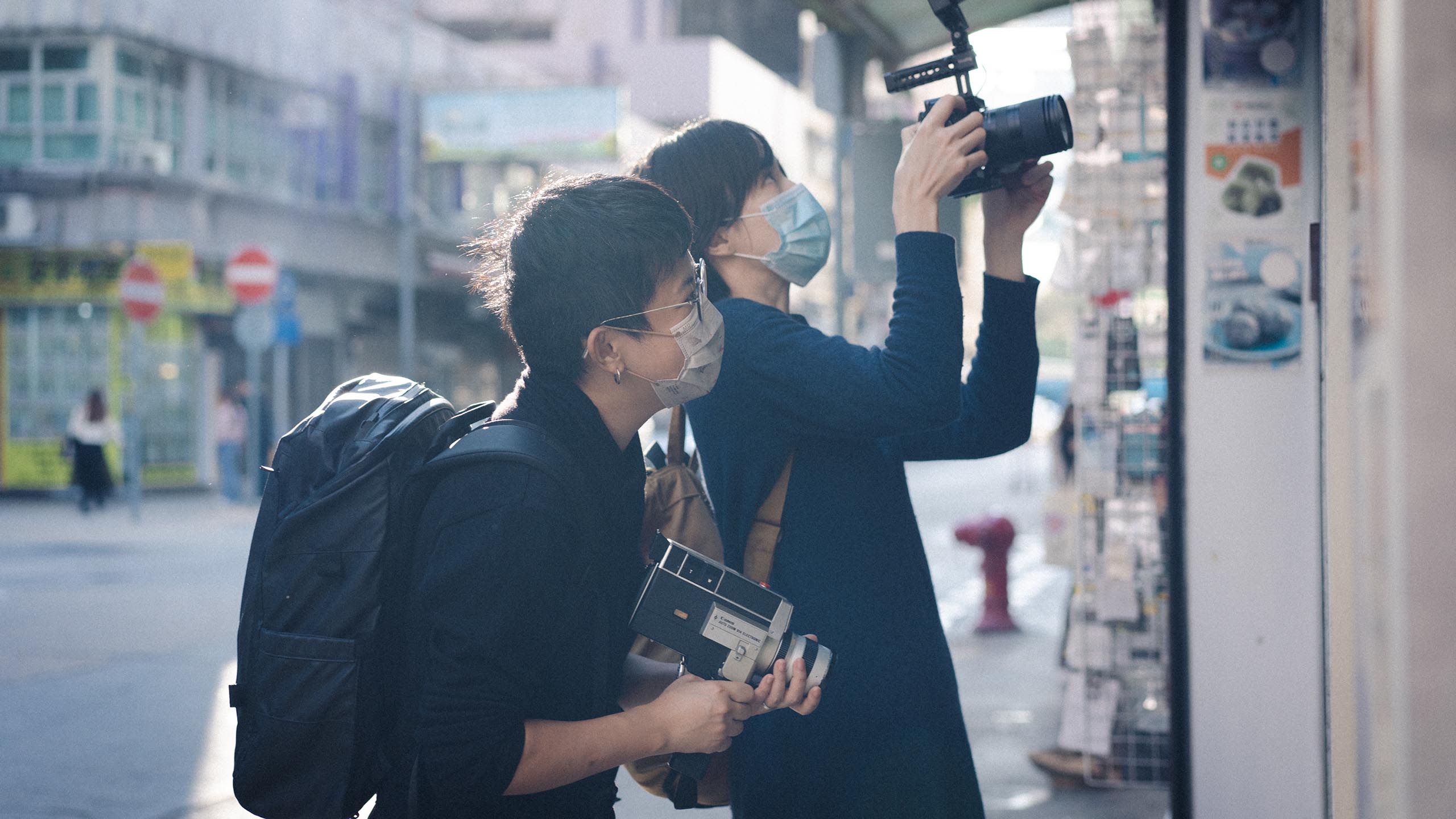 两位女性各自手执一部摄影机，在繁荣的香港社区中捕捉多变的街景。二人后方可见阳光。