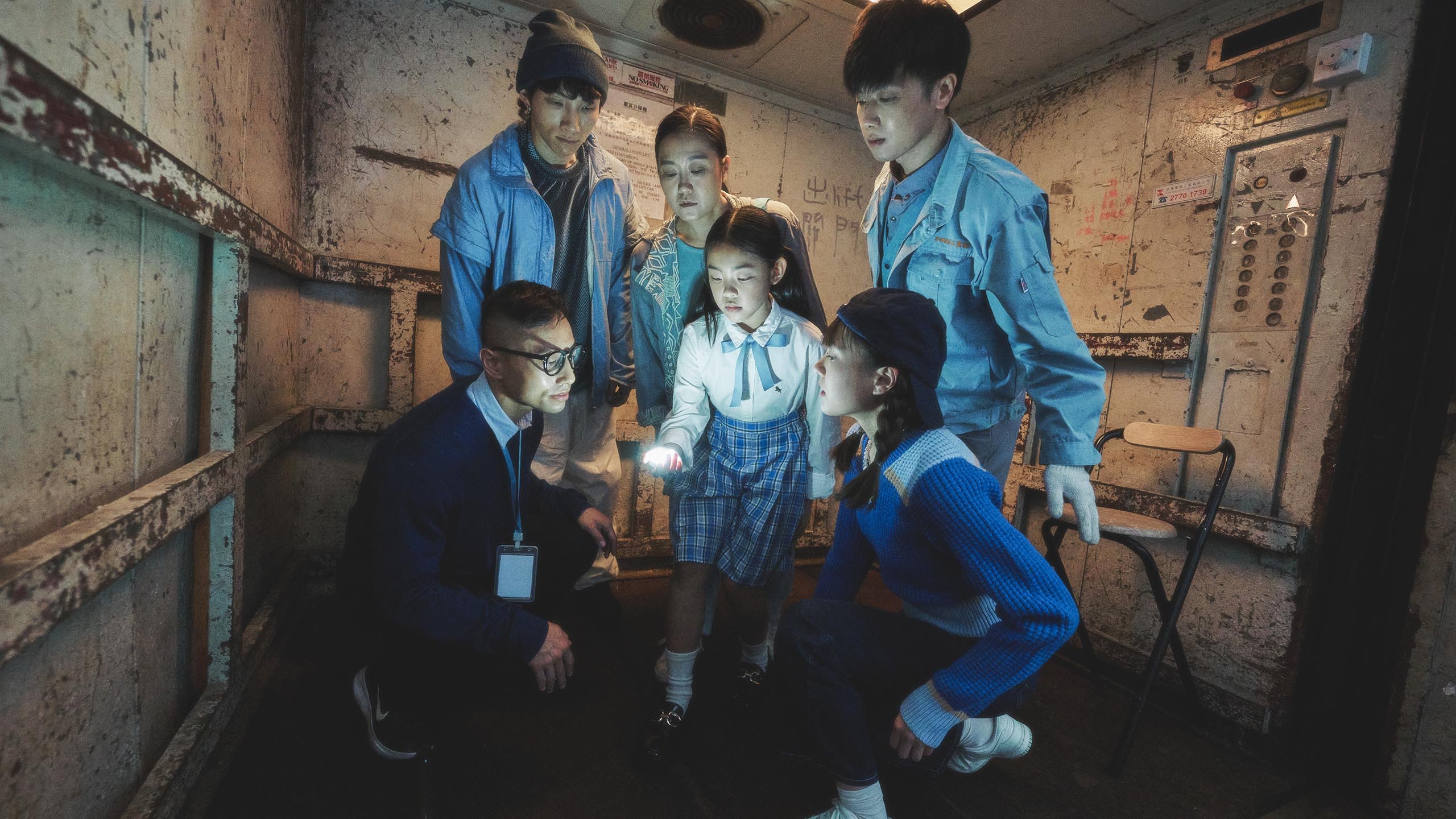 六位表演者聚在一台载货升降机中，当中身处众人中心的女孩拿着一个会发光的物体。