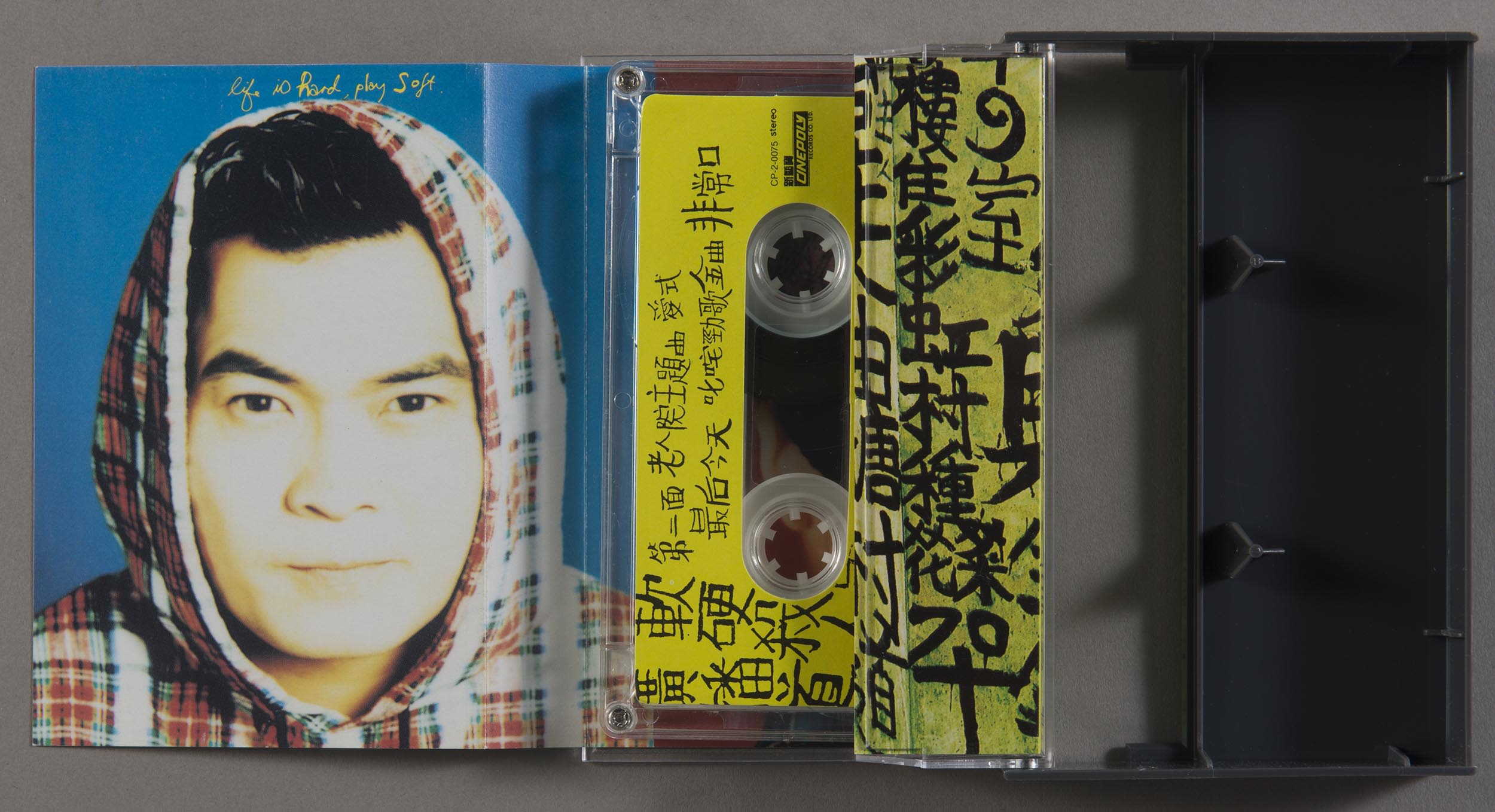 View of opened cassette box of Softhard’s Broadcast Drive Murder, with a photo of the singer Eric Kot wearing a plaid hoodie on the left. At the centre is the cassette tape, showing side b, which has a black, handwritten font on the yellow label. The encasing wrapper paper also has a similar graphic treatment.   