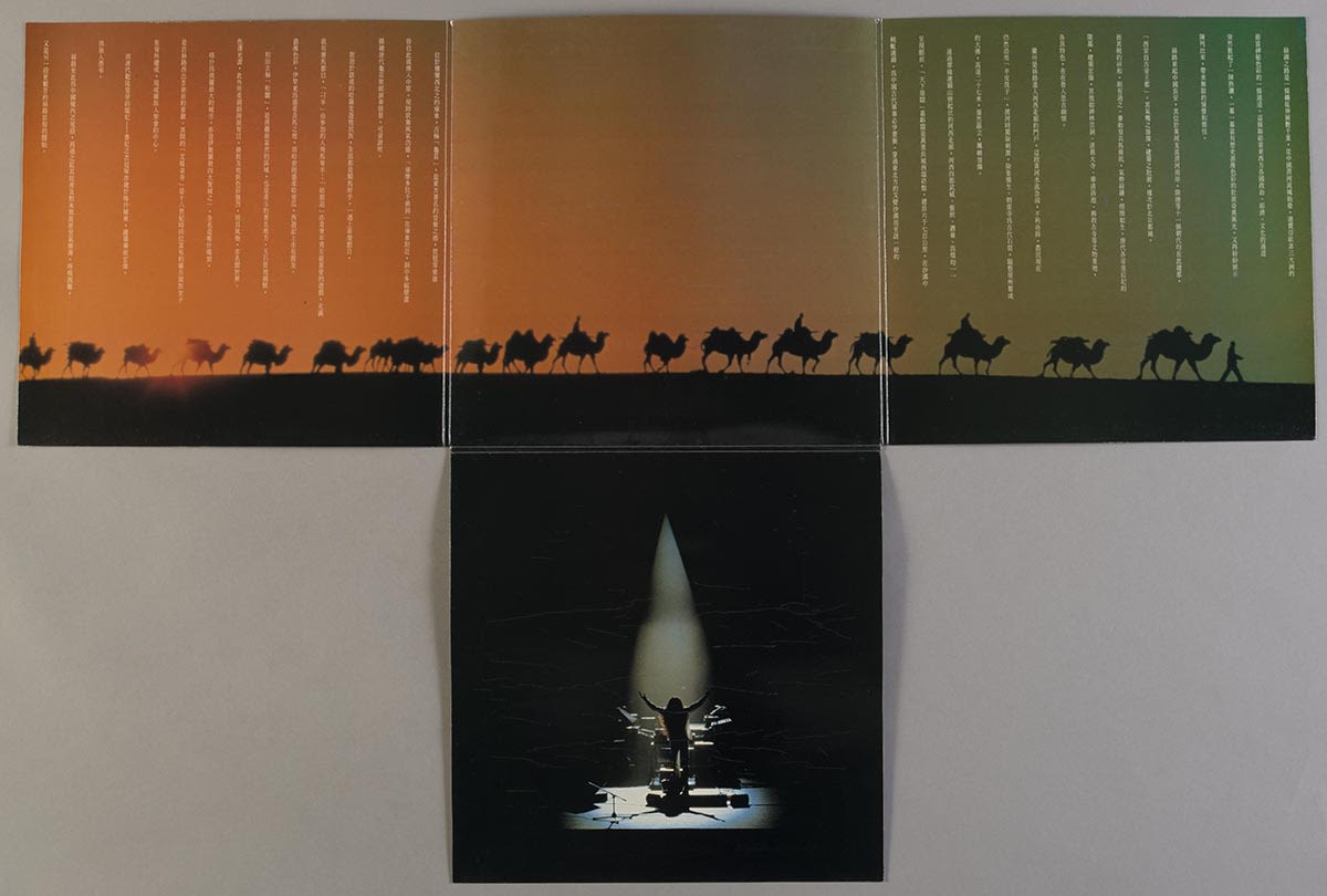 《丝绸之路巡礼名曲集》黑胶唱片封套的Ｔ字形四折内页。封套摊开后，可见一行旅人骑着骆驼横过沙漠的剪影在横向的三折内页上。从左到右，天色由绿色渐变成橙色，左右两页上有白色文字。中央下方的折页则有喜多郎演出时的背影。