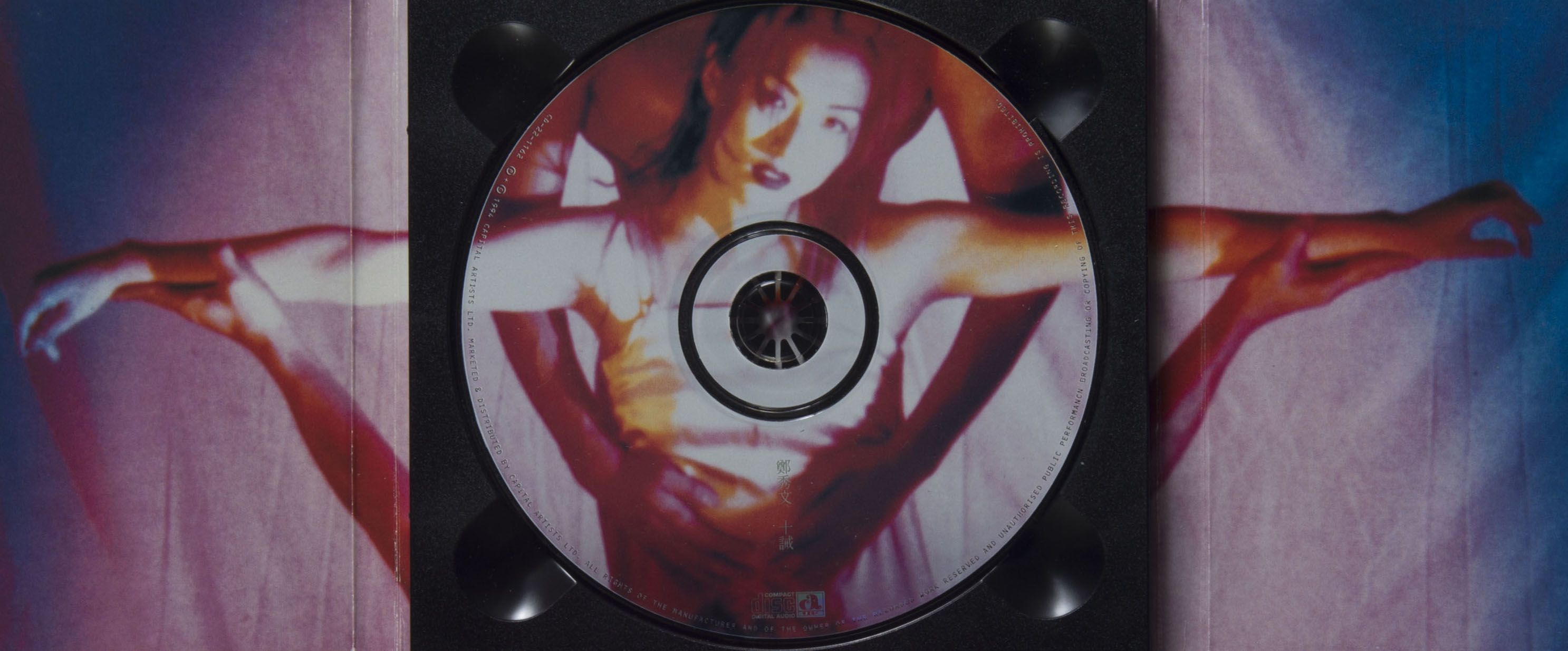 Interior of Sammi Cheng’s CD album Decalogue. Its image has an overall grainy effect, with yellow and magenta tones. On the disc is the singer in a strapped top, looking into the camera with her arms open. A male figure holds her waist from behind. Her right arm continues to extend beyond the disc into the left fold of the packaging.