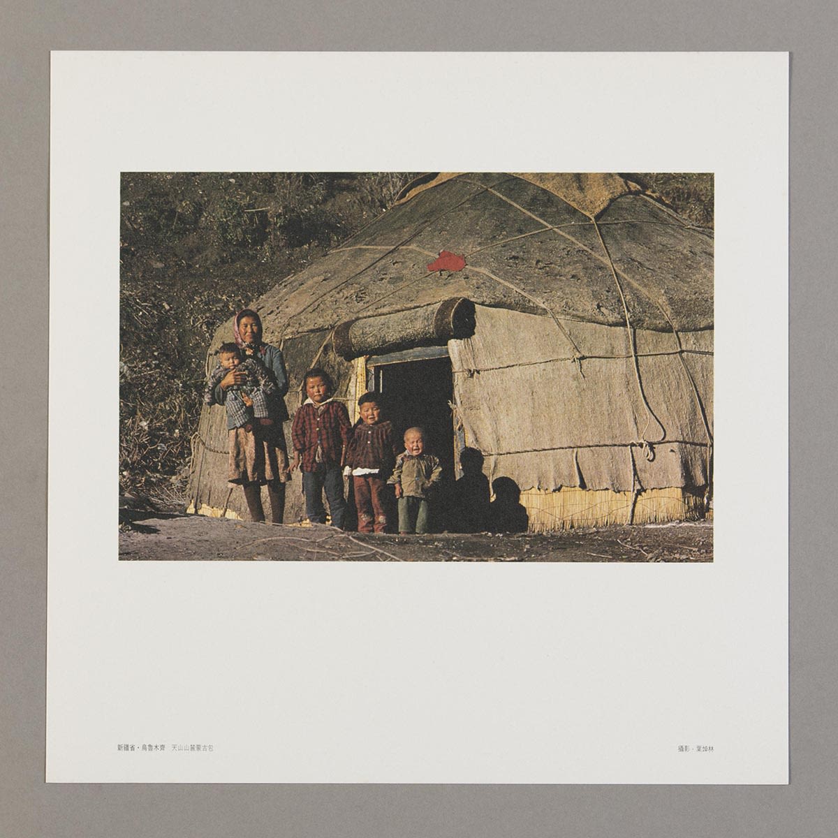 随《丝绸之路巡礼名曲集》黑胶唱片所附送的正方形明信片。白色边框内的照片可见手抱着小孩的母亲和三个由高至矮排列的孩子，他们站立在蒙古包前。照片摄于天山山脉一带。