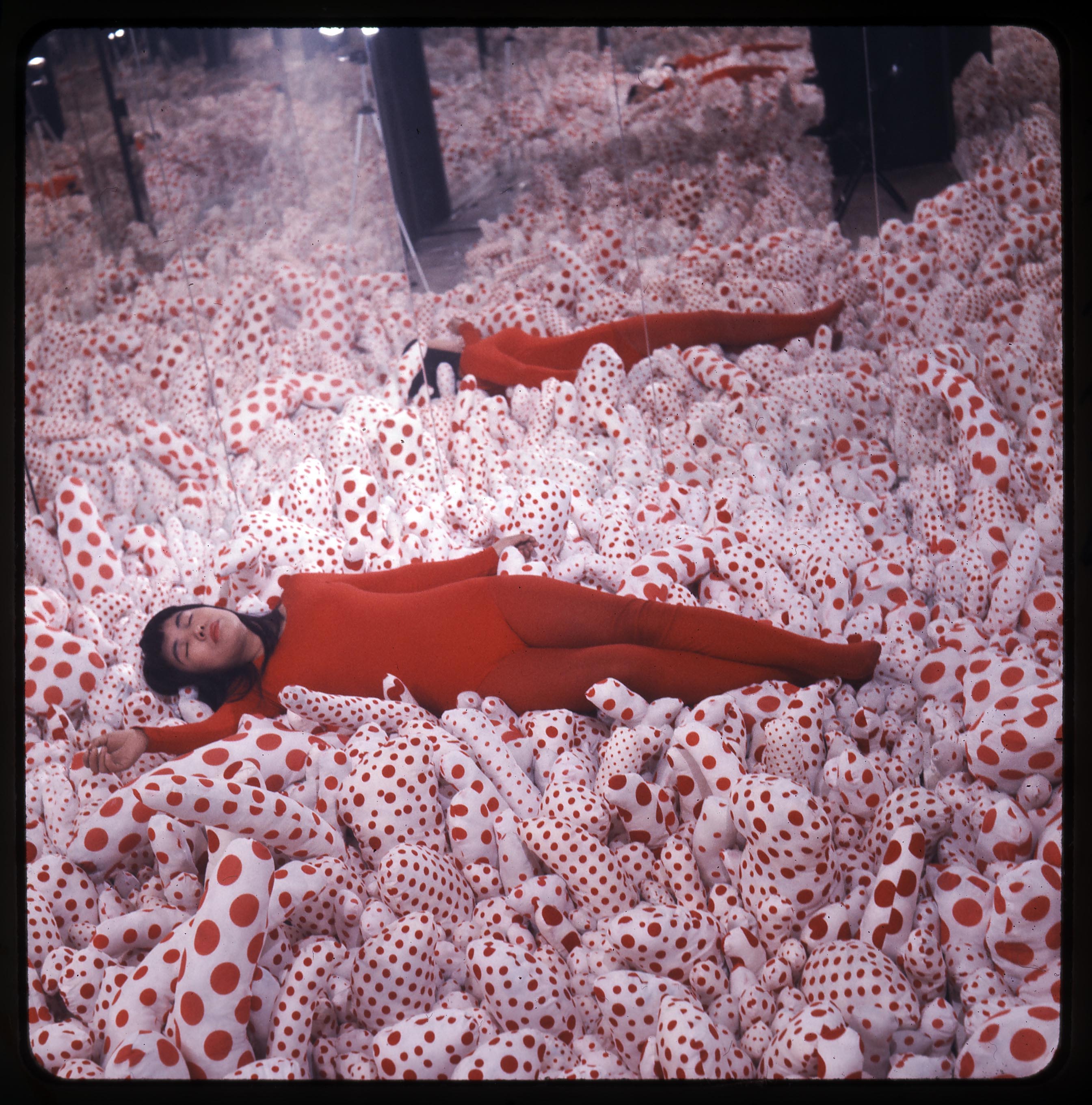 Photograph showing Kusama in a red bodysuit, lying on stuffed protrusions covered in red polka dots in a mirror room.