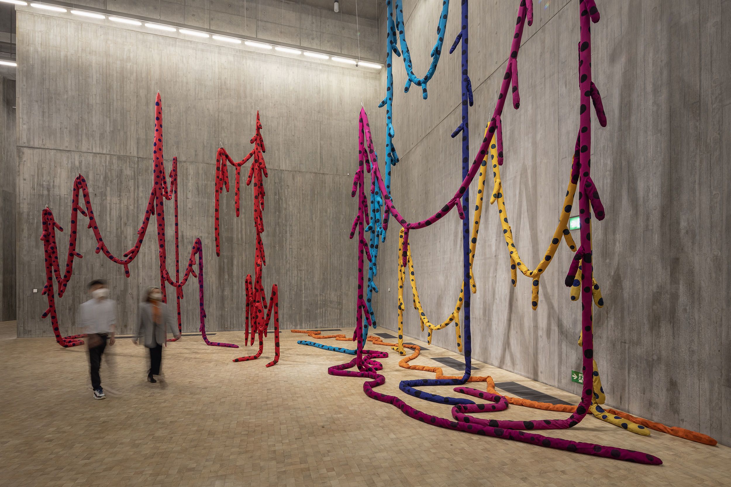 A soft sculpture featuring sewn tubes of suspended fabric in a cavernous concrete-walled exhibition space. The tubes are vibrant colours of red, purple, blue and yellow; black polka dots cover them. Two figures, blurred, walk past the sculpture.