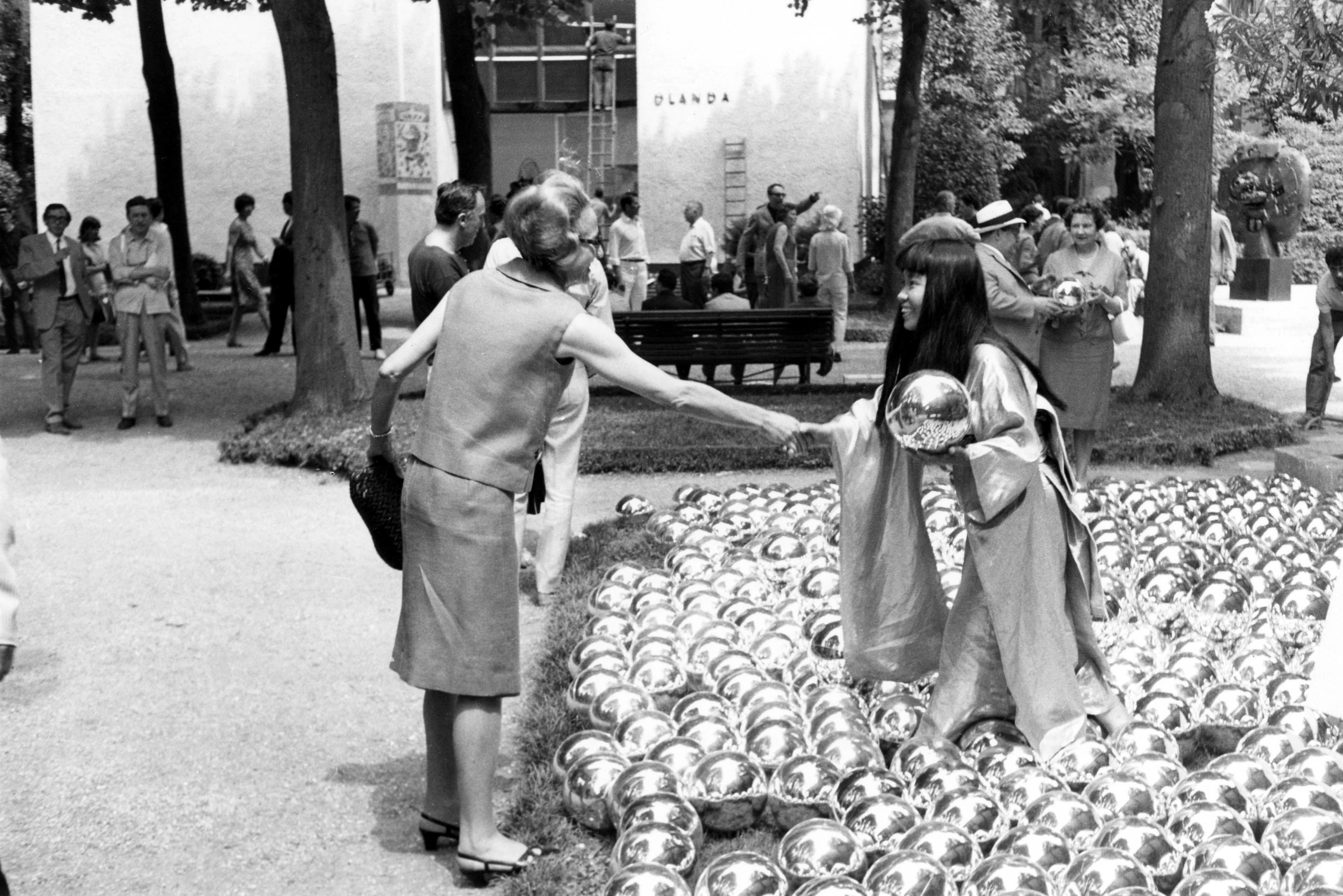 黑白照片中，草间弥生置身其作品《自恋庭园》，微笑着与一位参观者握手。艺术家站在大量镜球之间，并手持其中一个，似乎正在将它交给那位参观者。背景中另一位参观者的手中也捧着一个镜球。