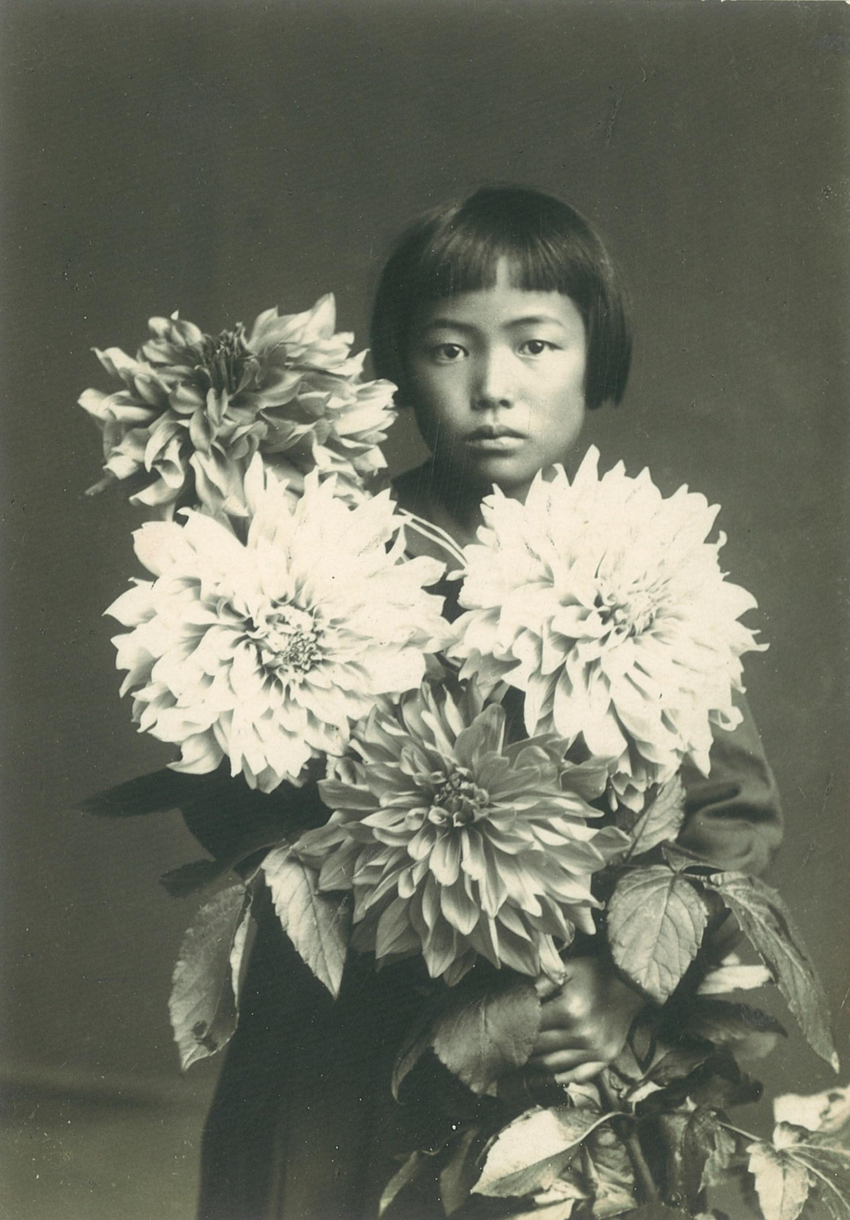 黑白肖像照片中，可见约十岁的草间弥生。她留着齐耳鲍伯短发，浏海短至眉毛上方，脸上没有明显的表情。她在照片中手执四朵盛开的大丽花。