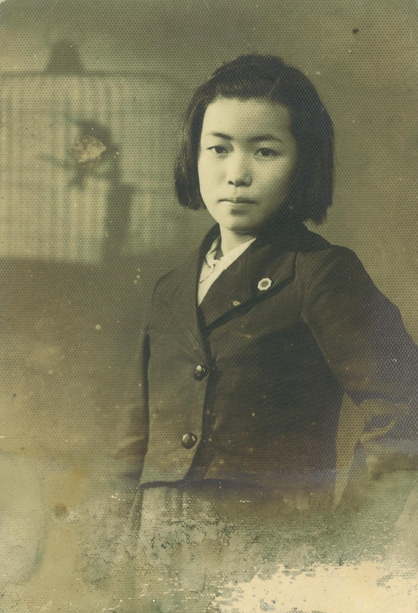 黑白肖像照片中，可见约十四岁的草间弥生。她穿着一件短版西装外套，一只手叉腰倾斜身体站立，外套的衣领上别着一枚徽章。背景中有一个鸟笼的影子。照片右下角已褪色。