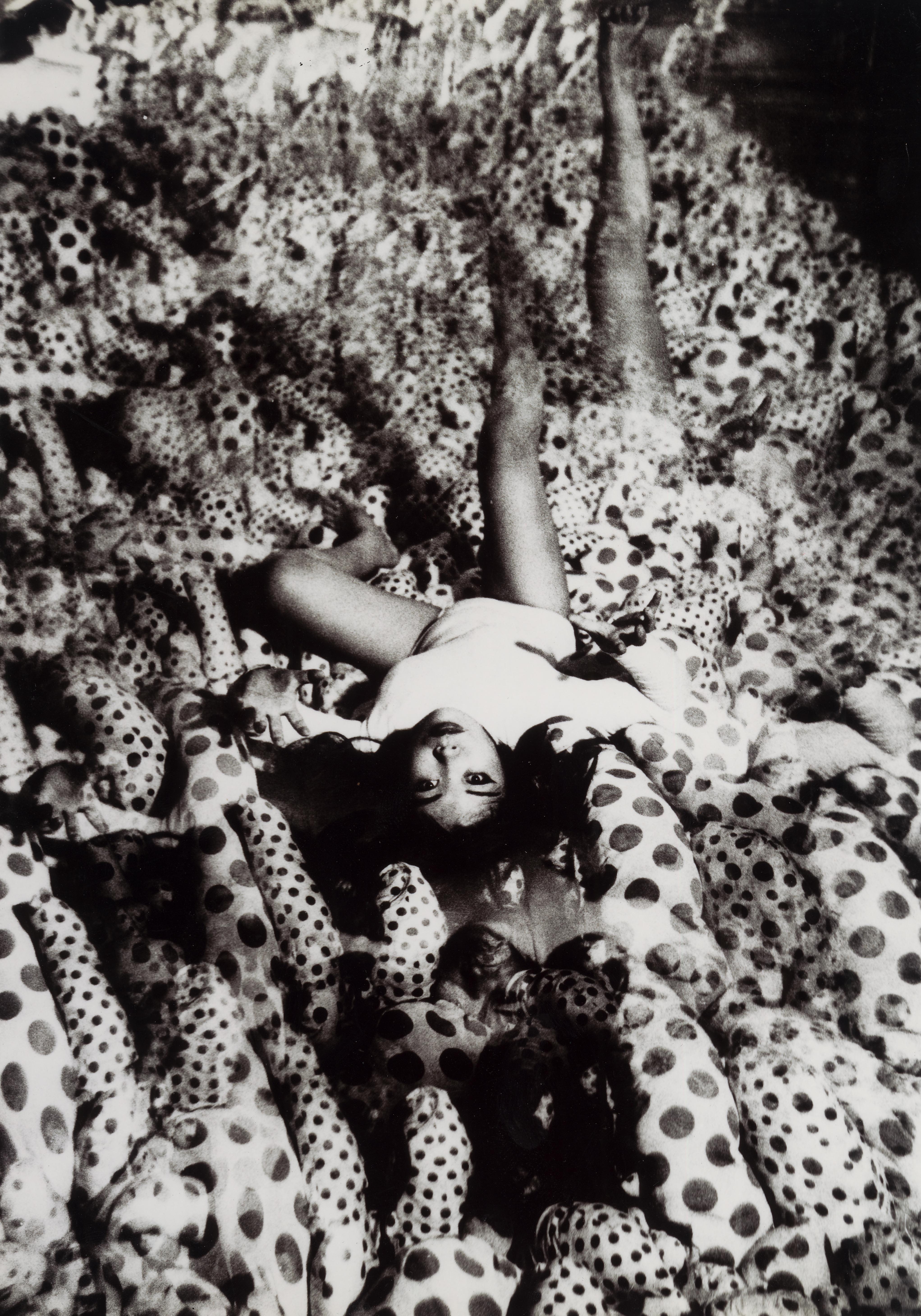 黑白照片摄于镜室，地面铺满带有圆点图案的突出状填充物，草间弥生手脚摊开躺在其上。