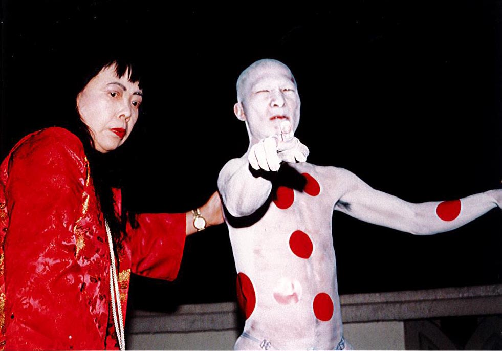 照片中呈现艺术展演的情景。身穿红衣的草间弥生将手放在表演者的背上，表演者身涂上红色圆点，他张开双臂，同时看着并指向镜头。