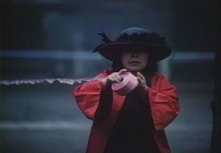 电影截图中的女子是艺术家草间弥生，她身穿一件飘逸的红色及膝长袍，头戴黑色宽边帽，手执一条长长的丝带。