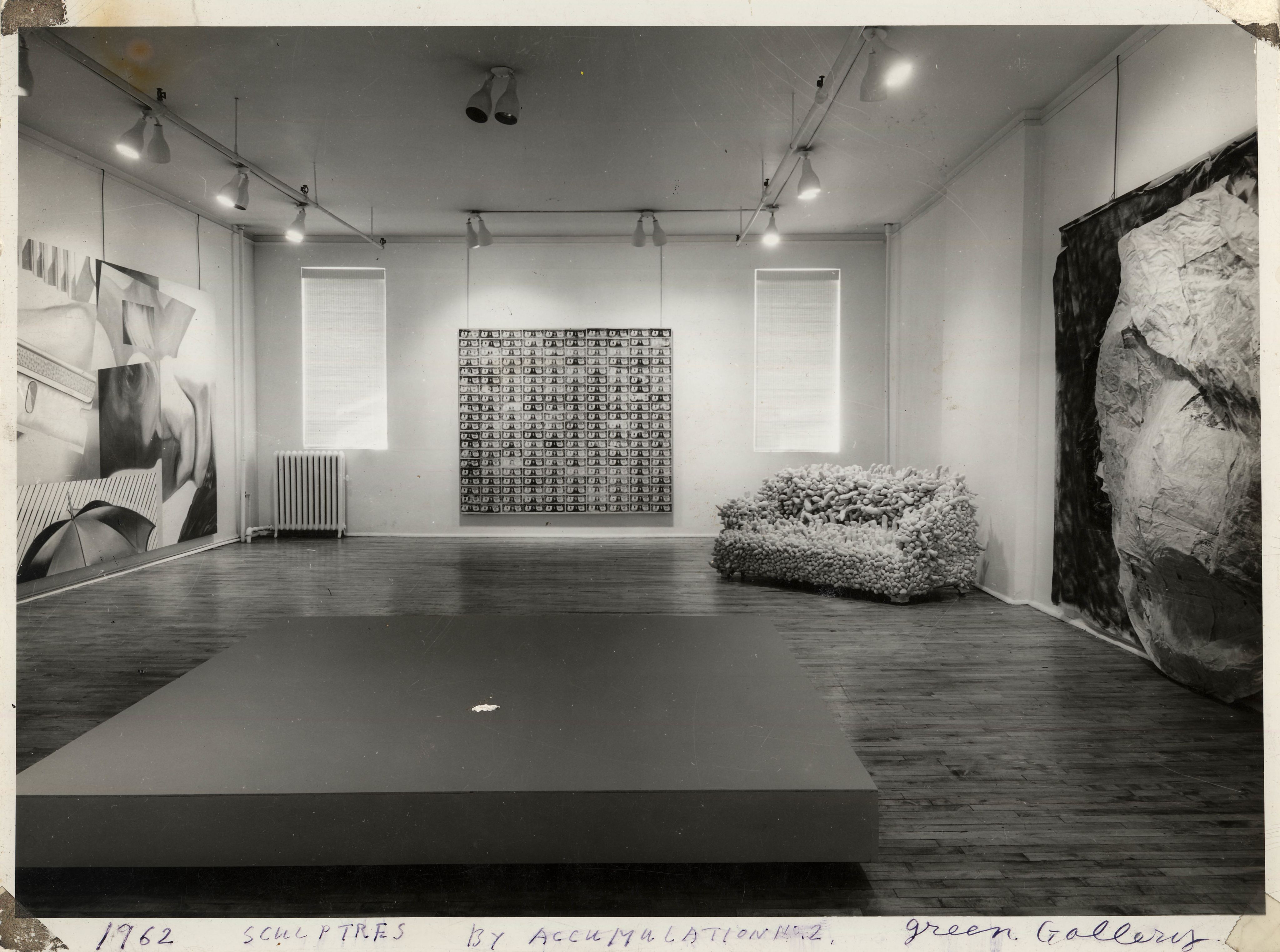 黑白照片中可见一个群展的展厅，当中陈列了多件雕塑和布本画作。右方一角摆放了一张双人沙发，表面覆满突出状的填充织物，沙发旁边挂着一幅由钞票组成的画作。左方墙上展示一幅由不同物件构成的作品。在前景中，一块深色的长方形厚板稍为升高。右方墙上挂着一块大布，表面依附着皱痕累累的大型囊状物。照片底部以蓝色墨水写上「1962年 积累而成的雕塑 2号，格林画廊」。