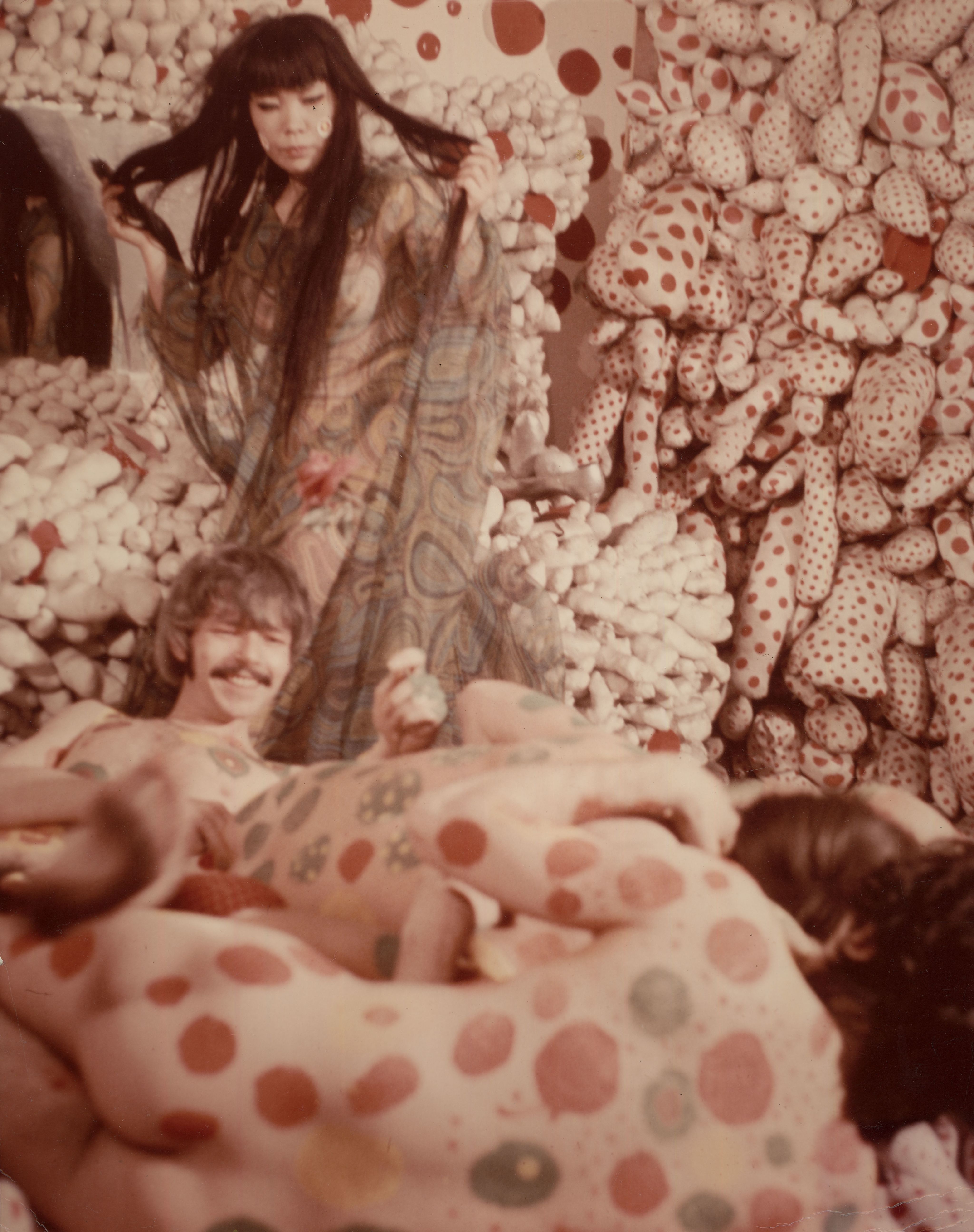 照片中是偶发艺术现场，房间内的墙壁覆盖着满布大小不一的突出状填充物，填充物上满布红色圆点。草间弥生披着半透薄纱，站着俯视三个或以上的人在地上纠缠，地上的人全部赤裸身躯，身上涂满圆点。