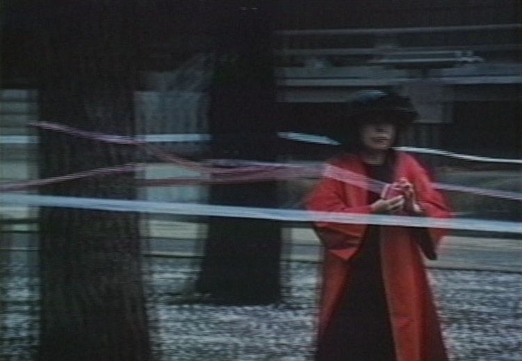 电影截图中的女子是艺术家草间弥生，她位于一个看似是公园的地方，身穿一件飘逸的红色及膝长袍，头戴黑色宽边帽，手执着一条长长的丝带，丝带绕着公园内几株树干。