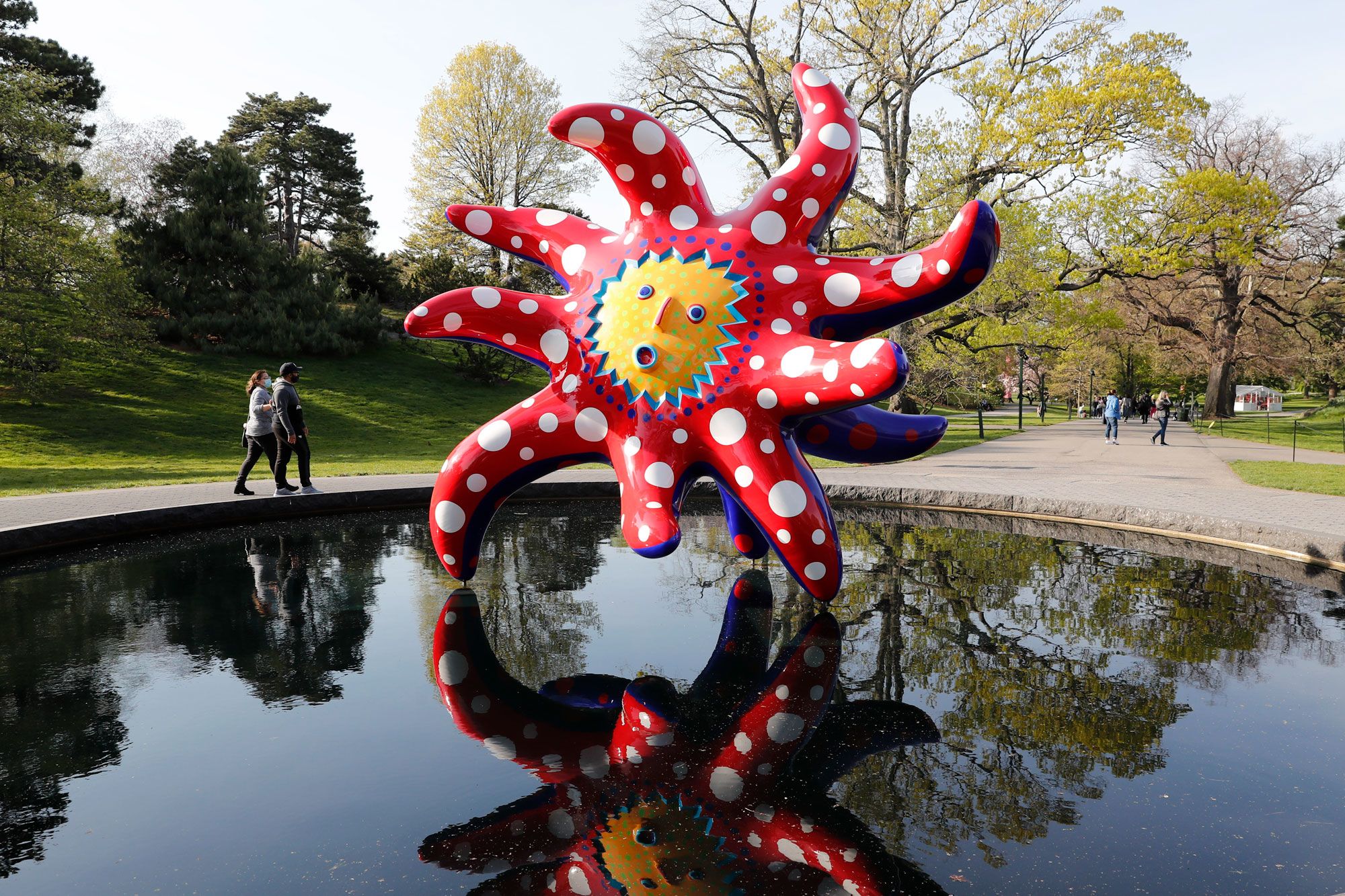 照片中的雕塑恍如太阳或花朵，竖立于公园的平静水面上。雕塑中央绘上一张卡通脸孔。背景中有树木和草地，还有两个在在水边走的行人。