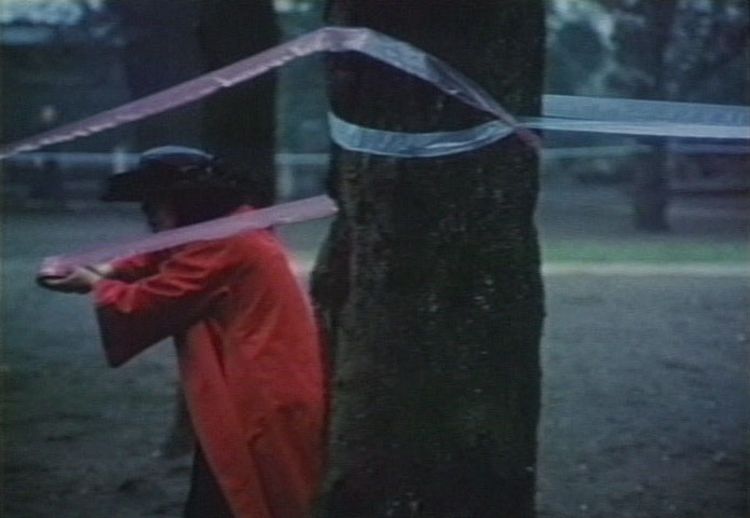 电影截图中的女子是艺术家草间弥生，她位于一个看似是公园的地方，身穿一件飘逸的红色及膝长袍，头戴黑色宽边帽，手上拉着一条长长的丝带，绕着公园内的几株树干。